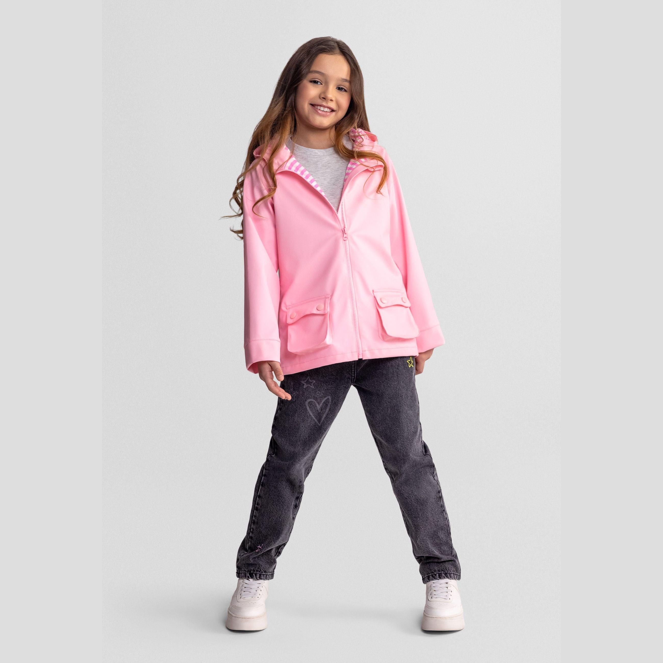 Girl wearing bright pink waterproof rain jacket with hood up, paired with dark grey jeans and white trainers, standing confidently against light grey studio background