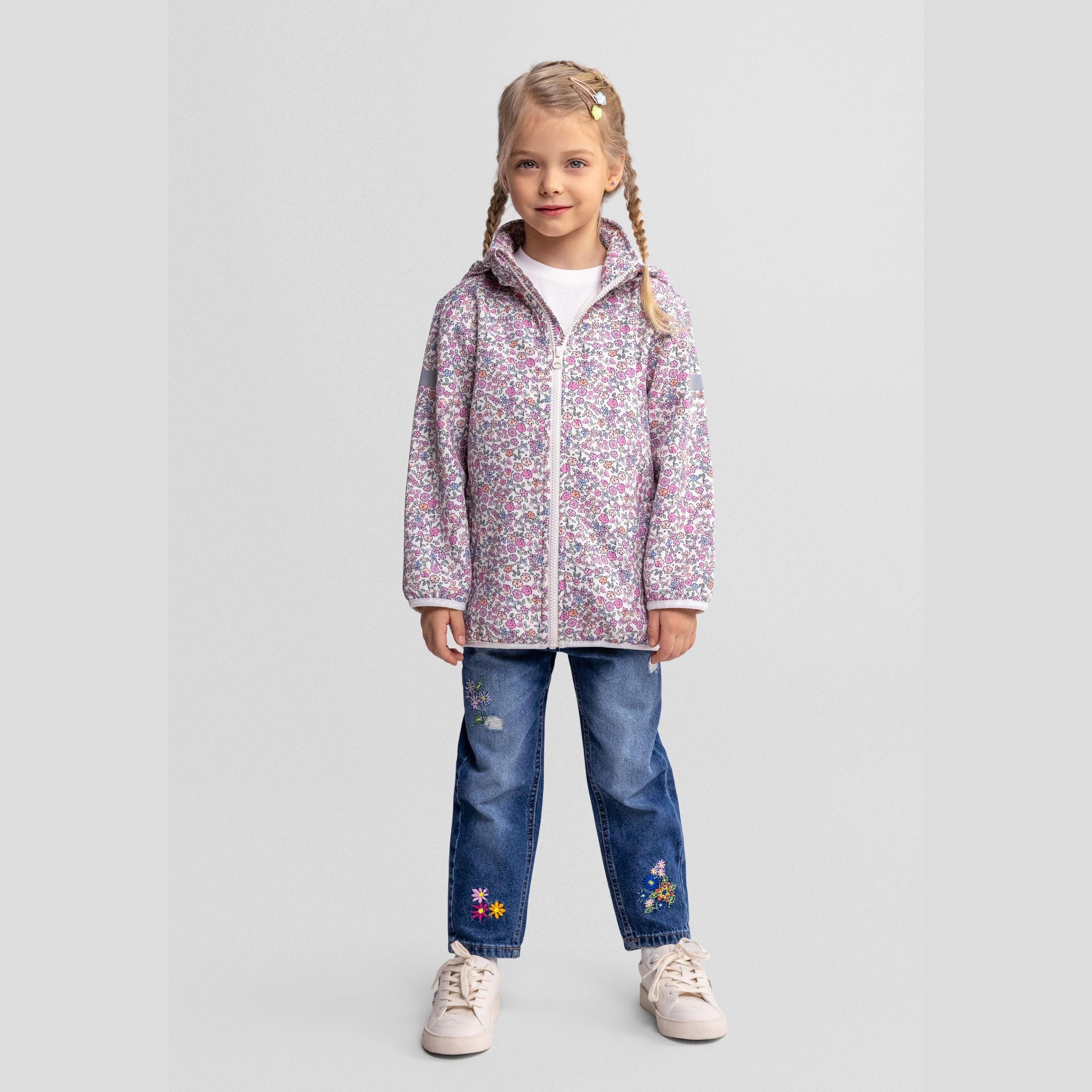 Young girl wearing off white softshell jacket with small floral print, blue jeans with embroidered flowers, standing against light grey background