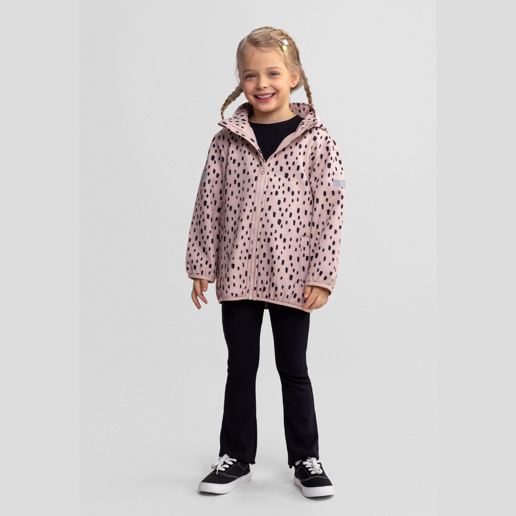 Young girl wearing pink softshell jacket with black spots over black outfit, standing against light grey background with blonde braided hair and yellow hair accessory