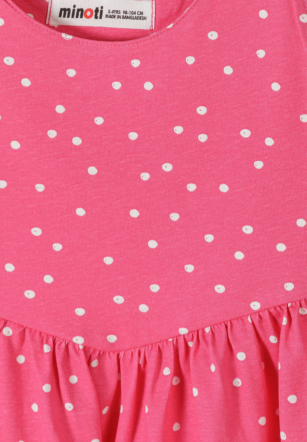 Close-up of the pink sundress's irregular white polka dots and gathered waist seam, with a Minoti neck label.
