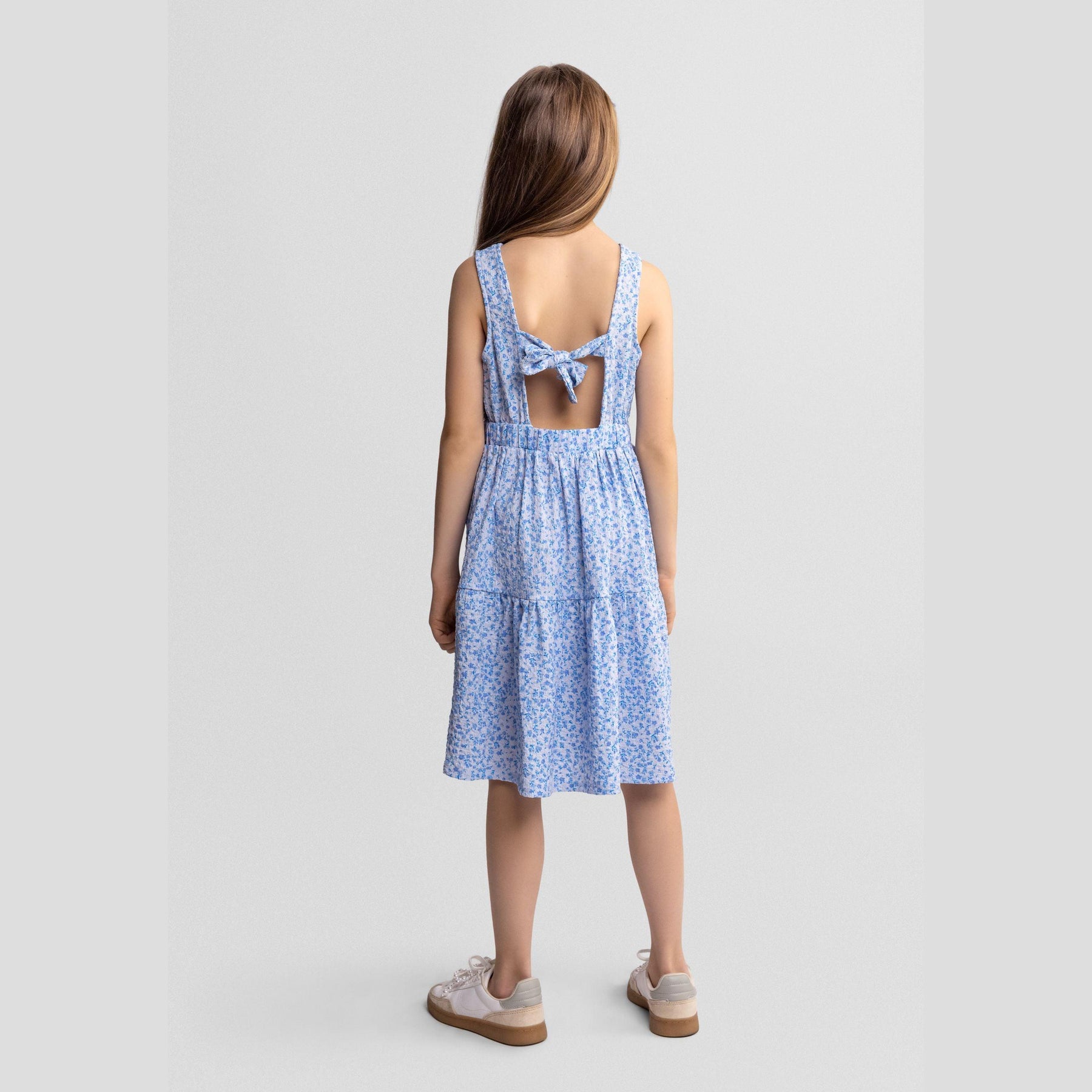 Back view of girl wearing blue floral tiered dress with bow tie detail and beige sandals on white background