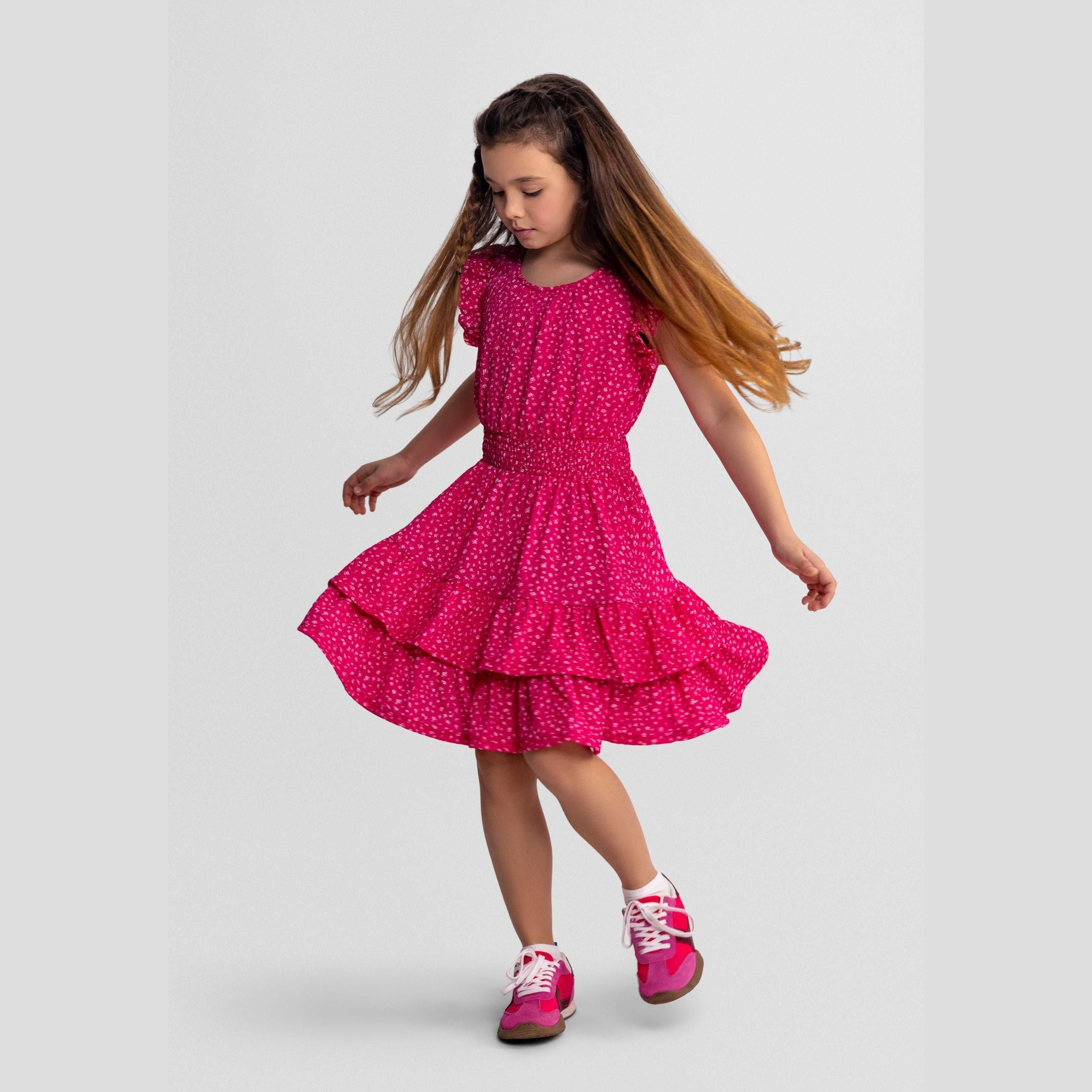 Young girl wearing vibrant pink floral dress with frill sleeves and tiered skirt, dancing against white background, showing dress movement and fit