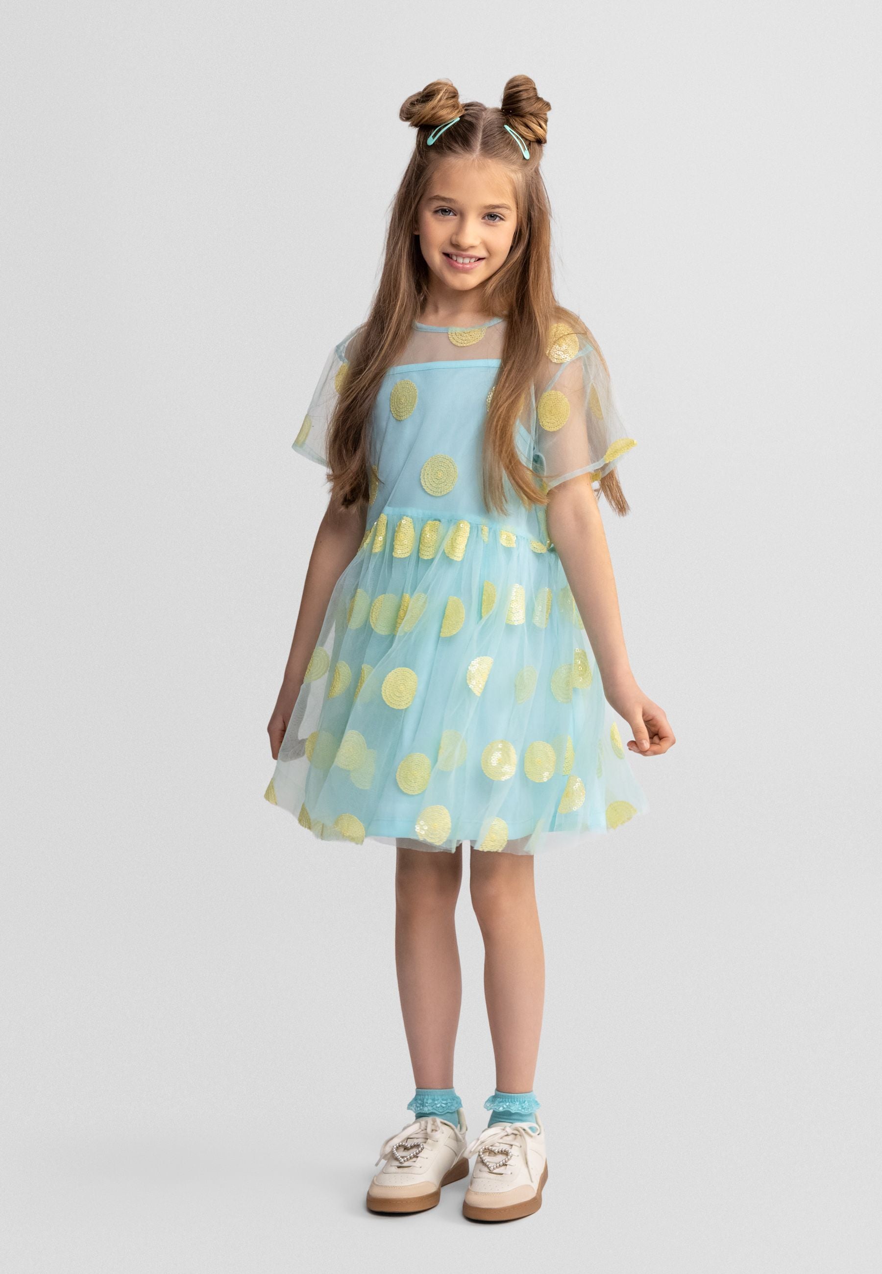 Young girl wearing turquoise tulle party dress with gold sequin polka dots, short sleeves, layered skirt, standing against white studio background