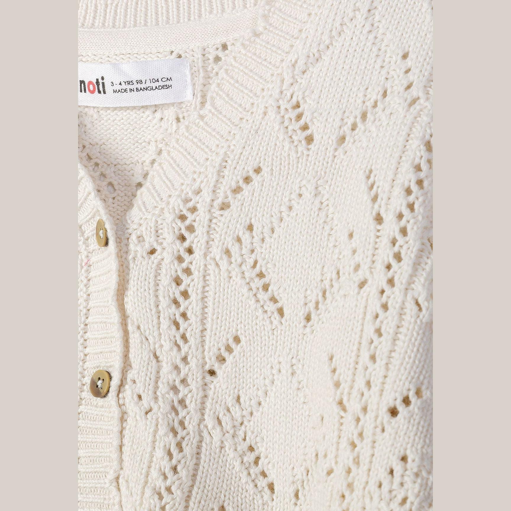 Close-up detail of white pointelle cardigan showing textured knit pattern, wooden buttons and Minoti care label against neutral background