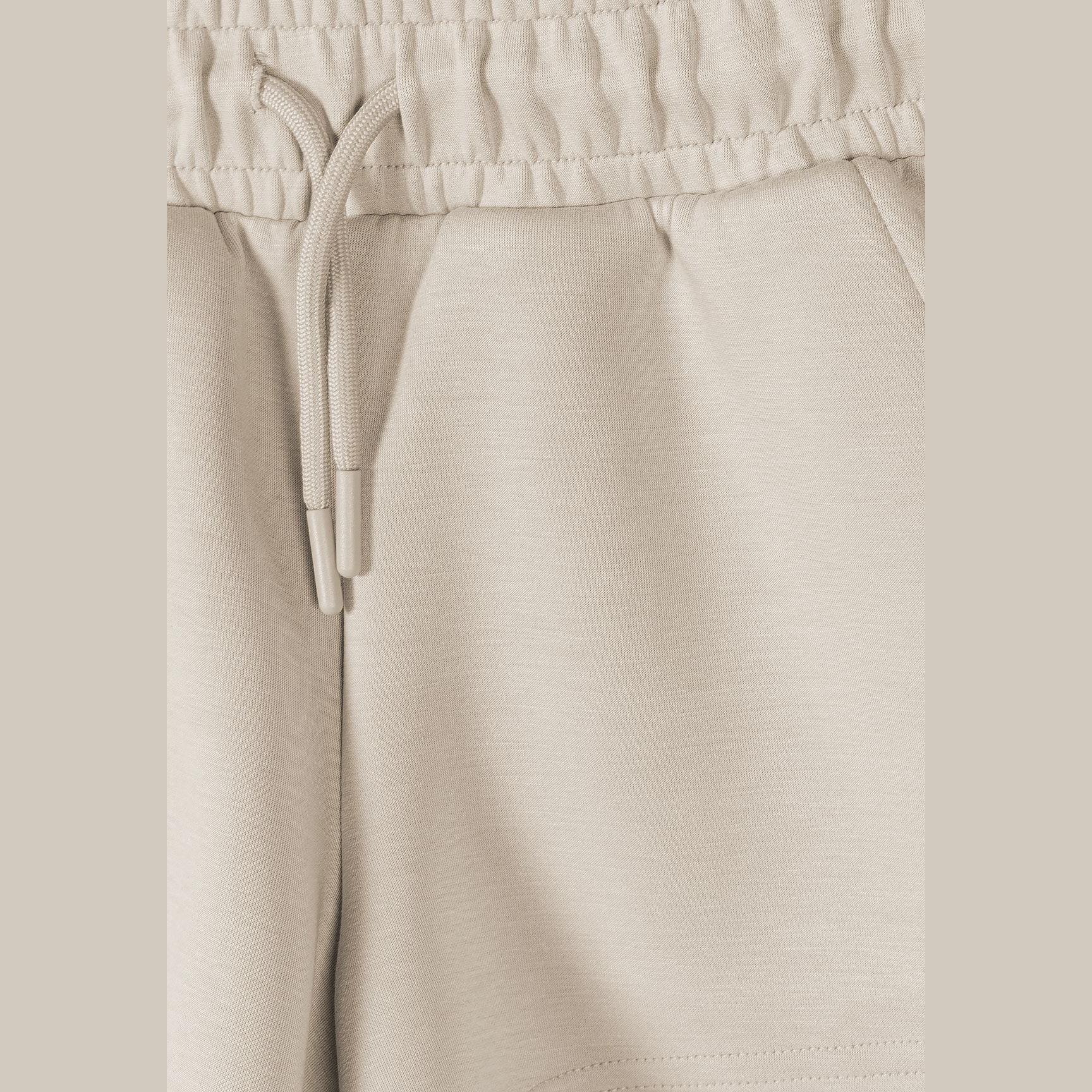 Close-up detail of beige shorts showing elasticated waistband with drawstring ties on neutral background
