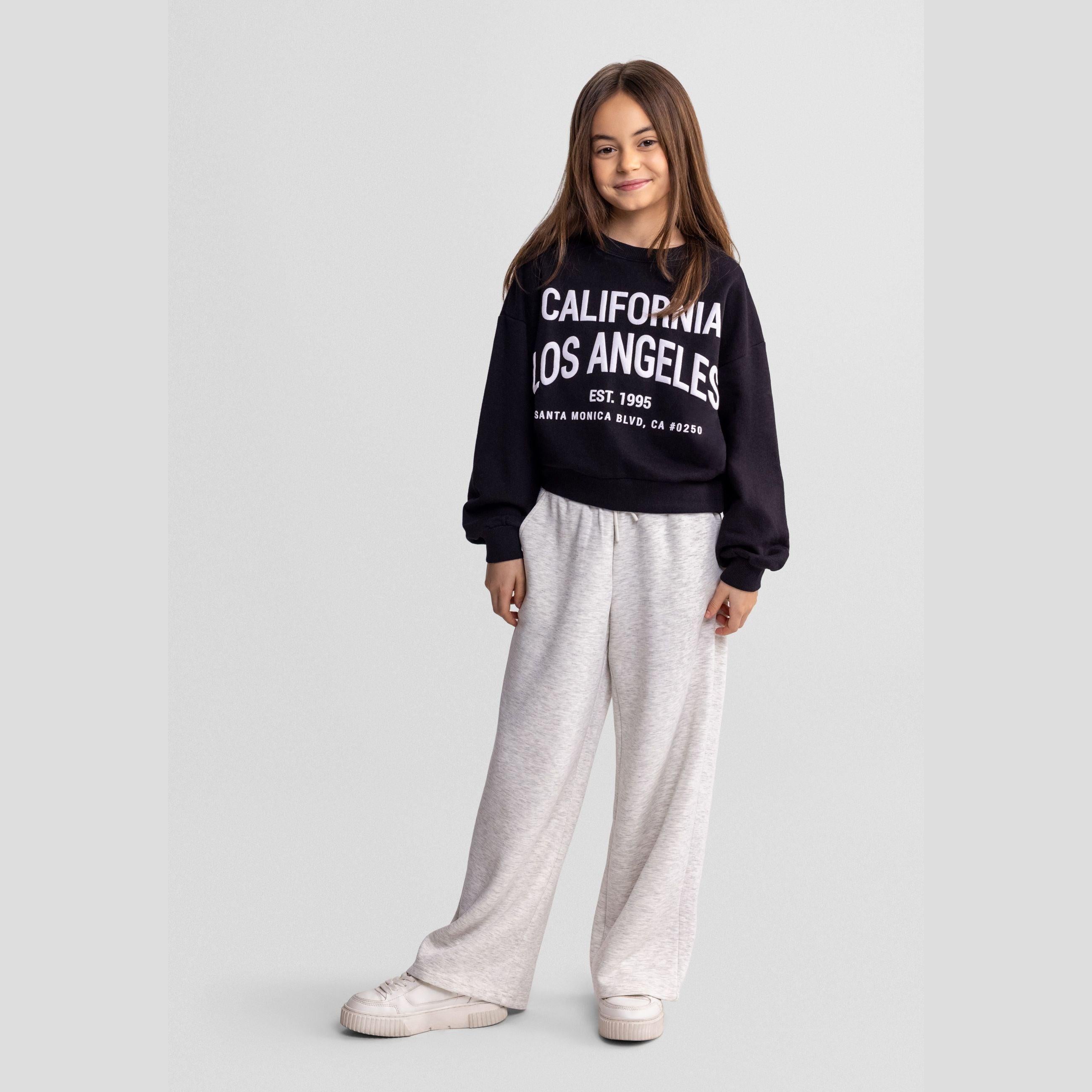 Young girl wearing wide leg mottled grey joggers with navy California Los Angeles sweatshirt, standing against light grey background