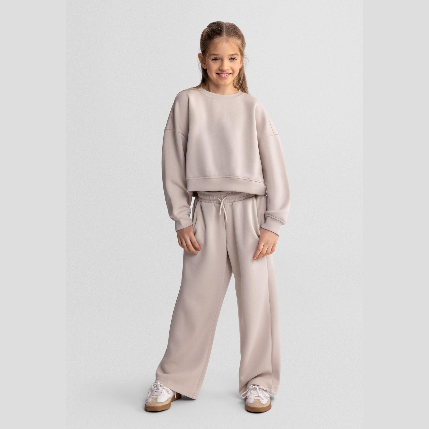 Girl wearing beige wide leg joggers with matching sweatshirt, standing against white background, showing full outfit coordination