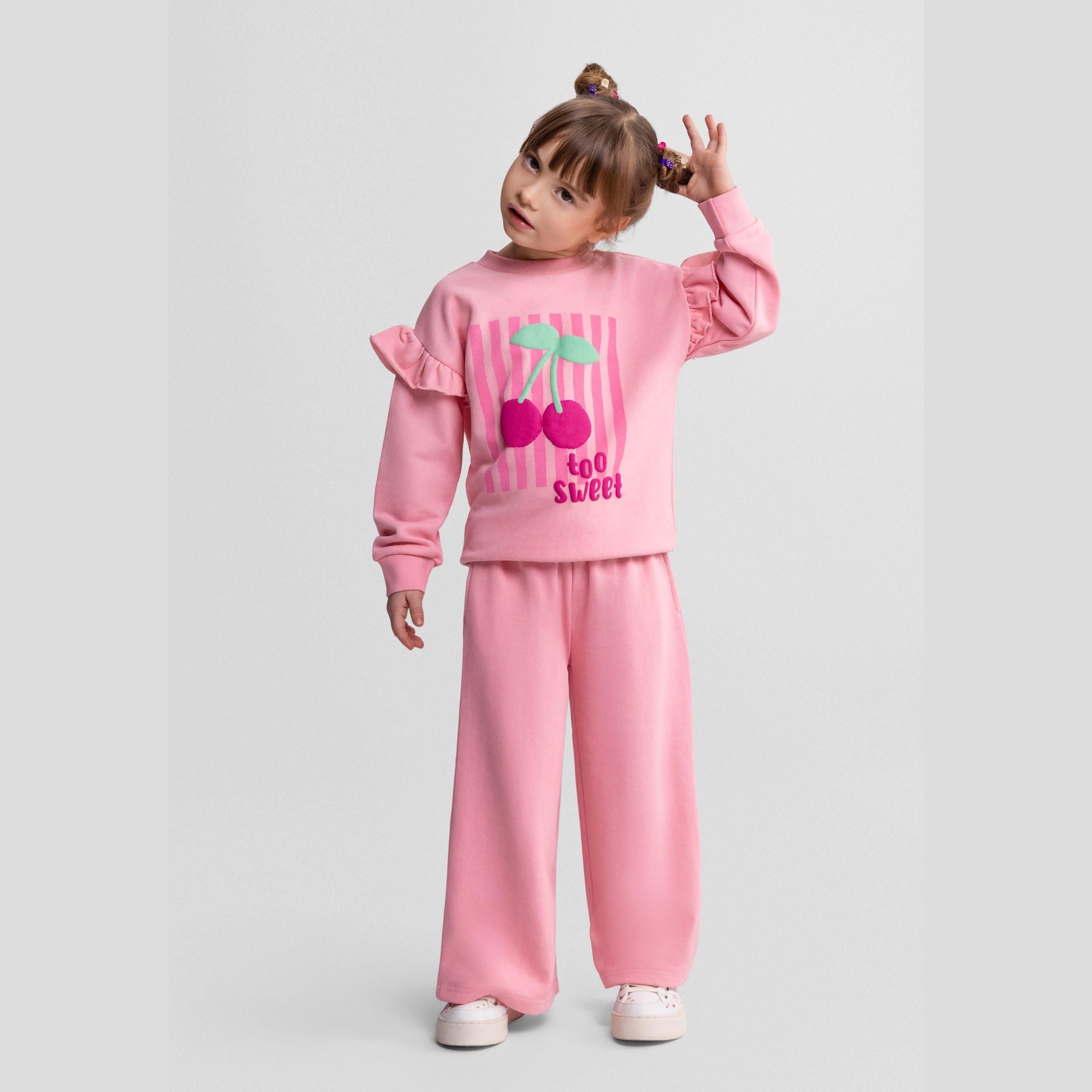 Young girl wearing pink cherry tracksuit with ruffle sleeves, posing playfully against white studio background