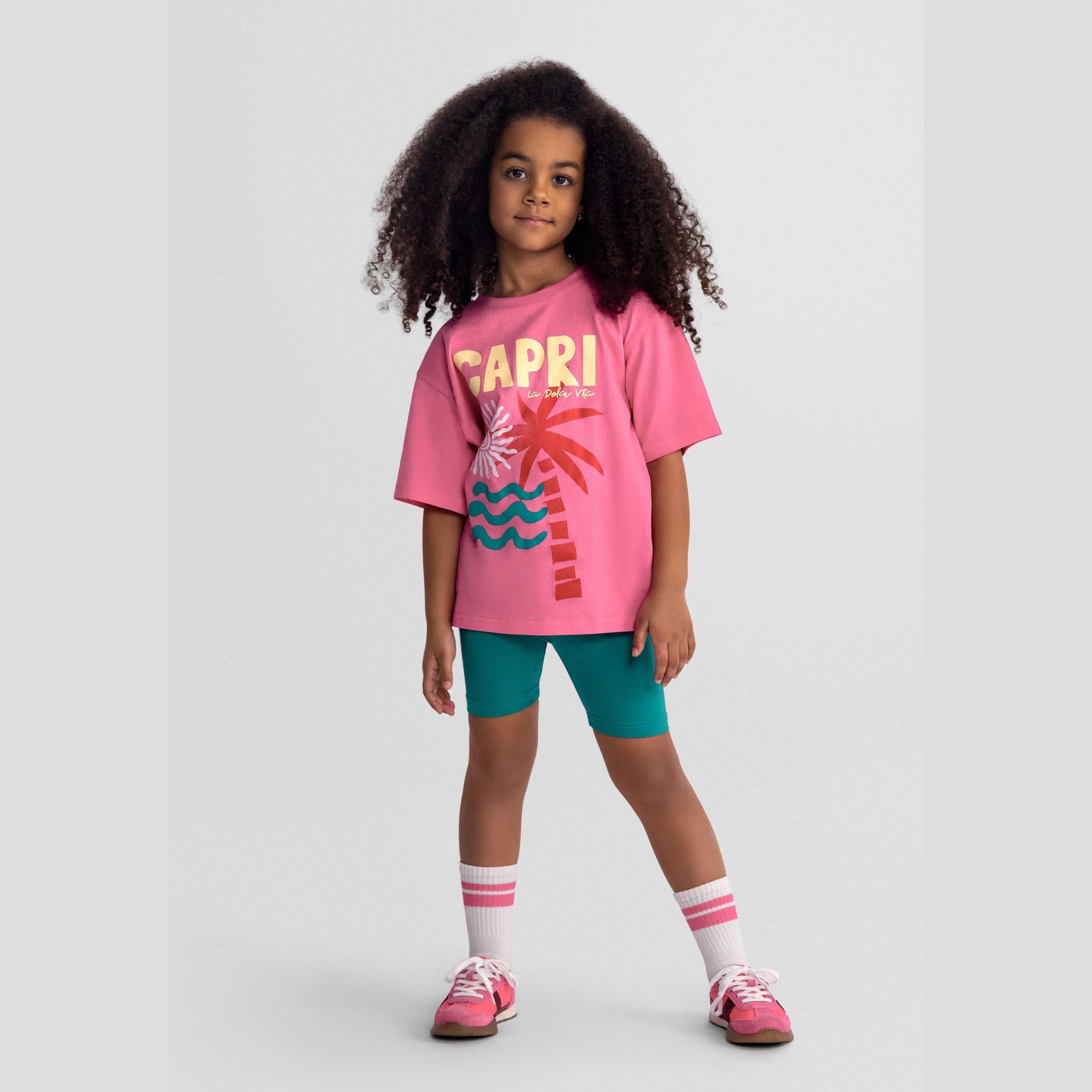 Young girl wearing pink oversized t-shirt with CAPRI text and palm tree graphic paired with teal cycling shorts, white striped socks and pink sneakers on grey background