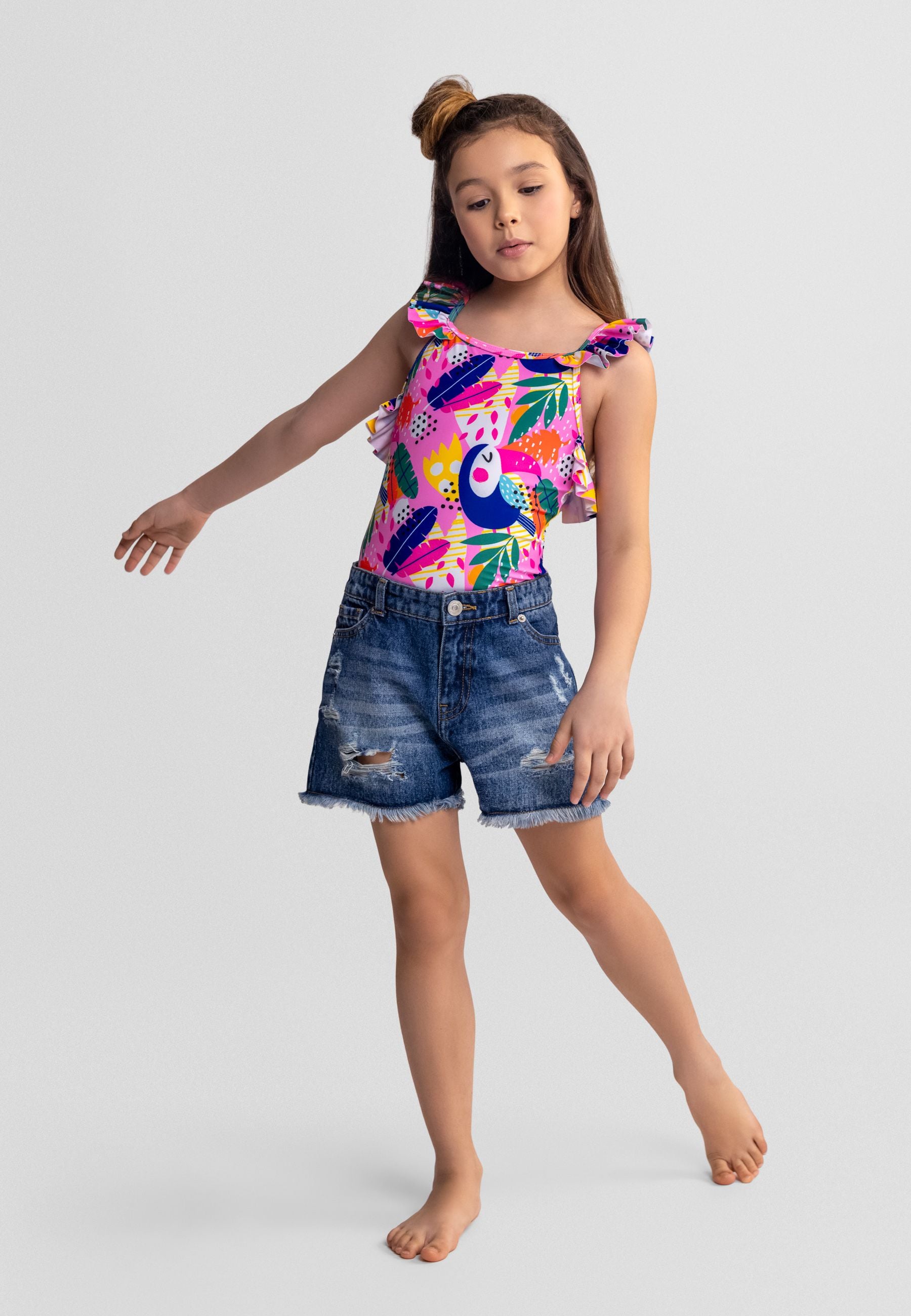 Young model wearing tropical print bodysuit with toucan design paired with light blue distressed denim shorts, standing against white studio background