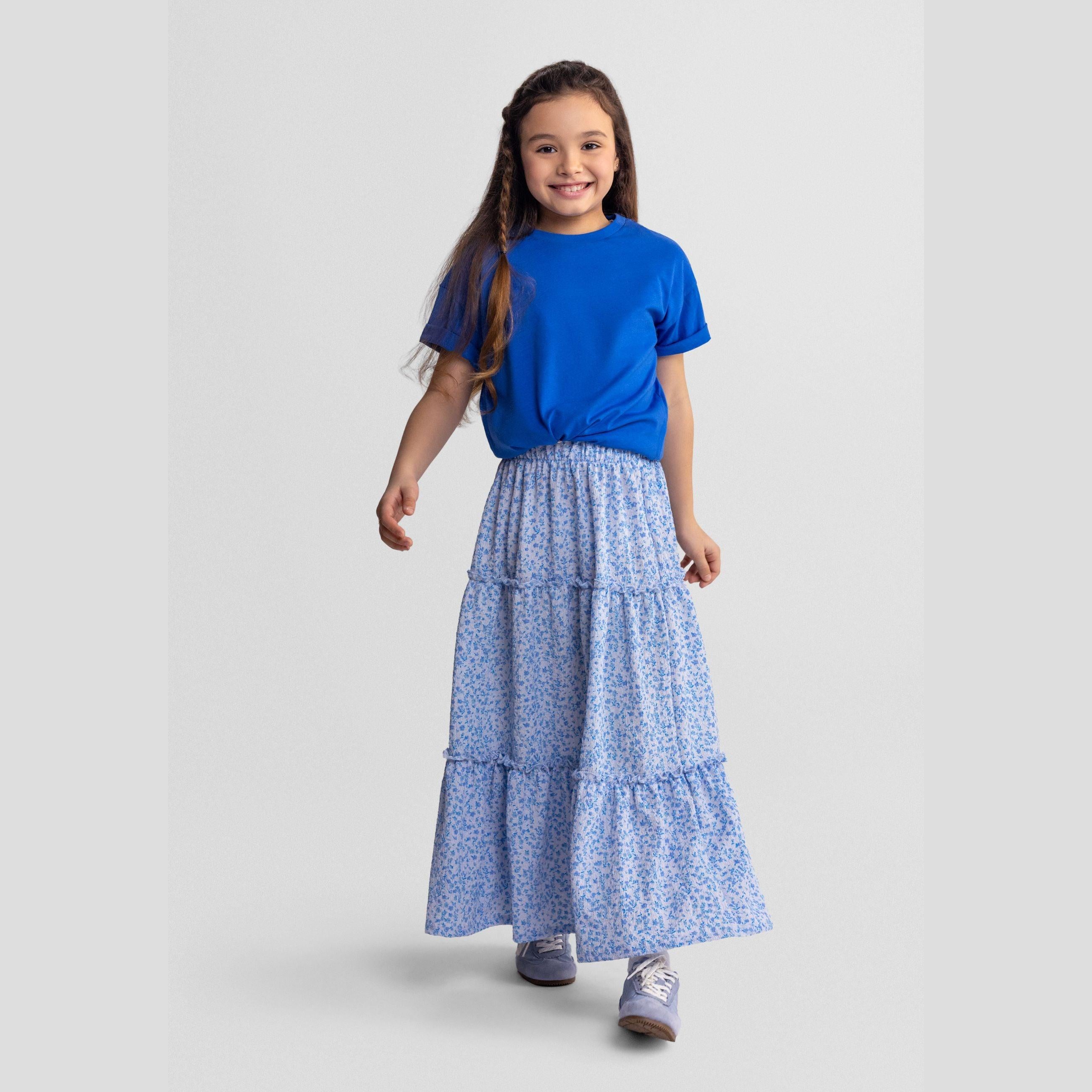 Young girl wearing blue t-shirt and blue floral tiered maxi skirt, standing against light grey background
