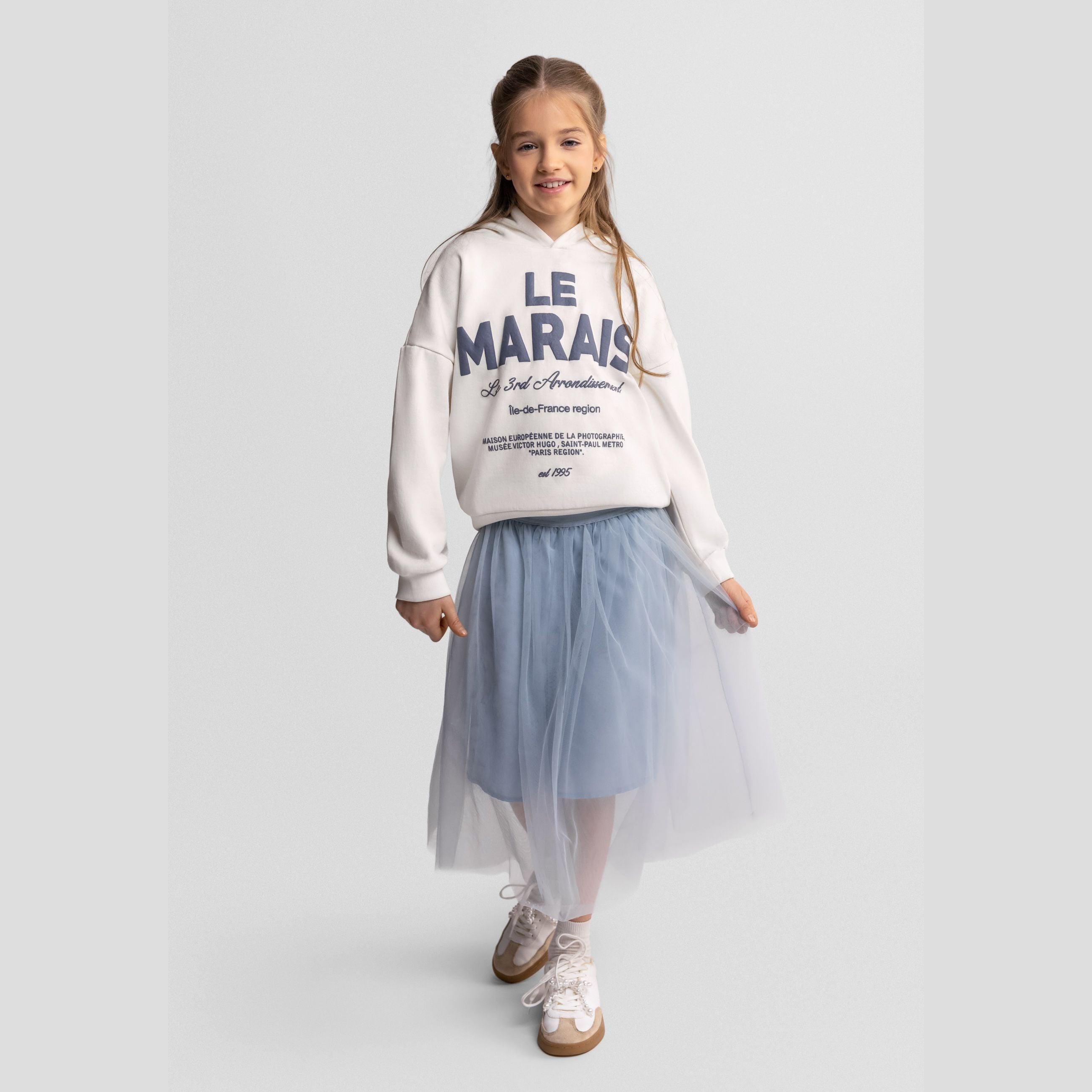 Young girl wearing grey pleated tulle maxi skirt with white hoodie, standing against white background, full body shot showing the flowing movement of the skirt