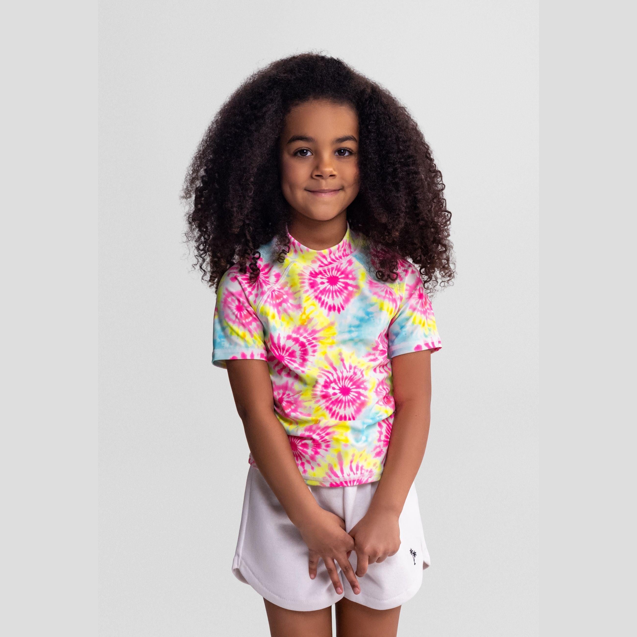 Young girl wearing colourful tie-dye swim t-shirt with spiral patterns in pink, yellow and blue, paired with white shorts, standing against light grey background