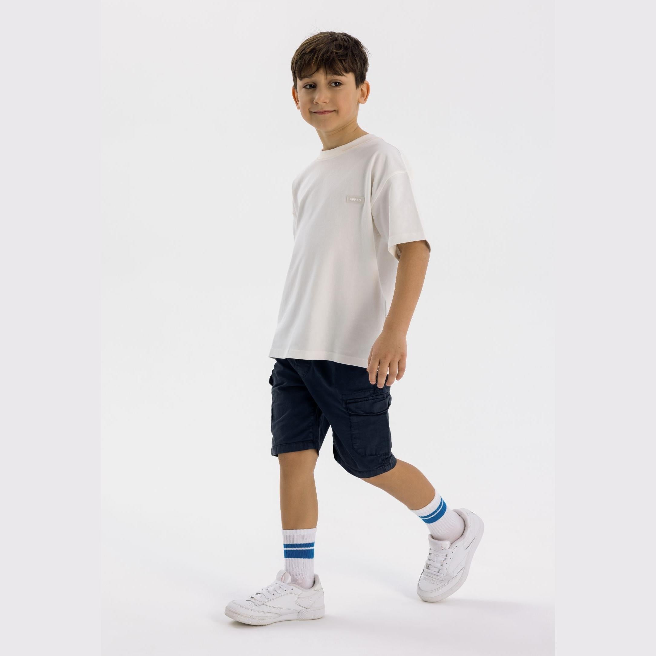 Young boy wearing white t-shirt and navy combat shorts with cargo pockets, styled with white sneakers and striped socks against white background