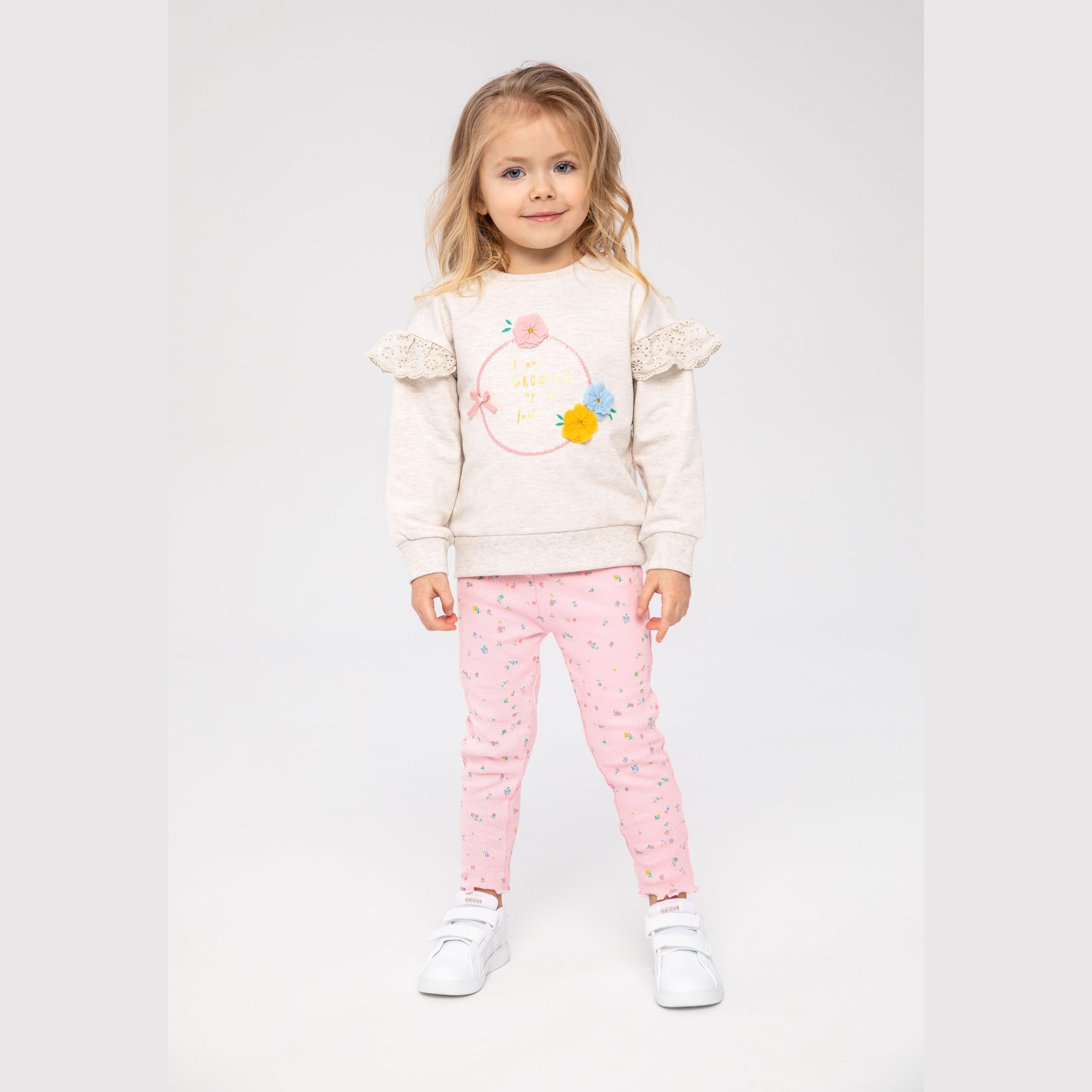 Young blonde girl wearing beige fleece sweatshirt with frill sleeves and 3D flower applique, paired with pink ribbed leggings with scattered pattern, standing against white studio background
