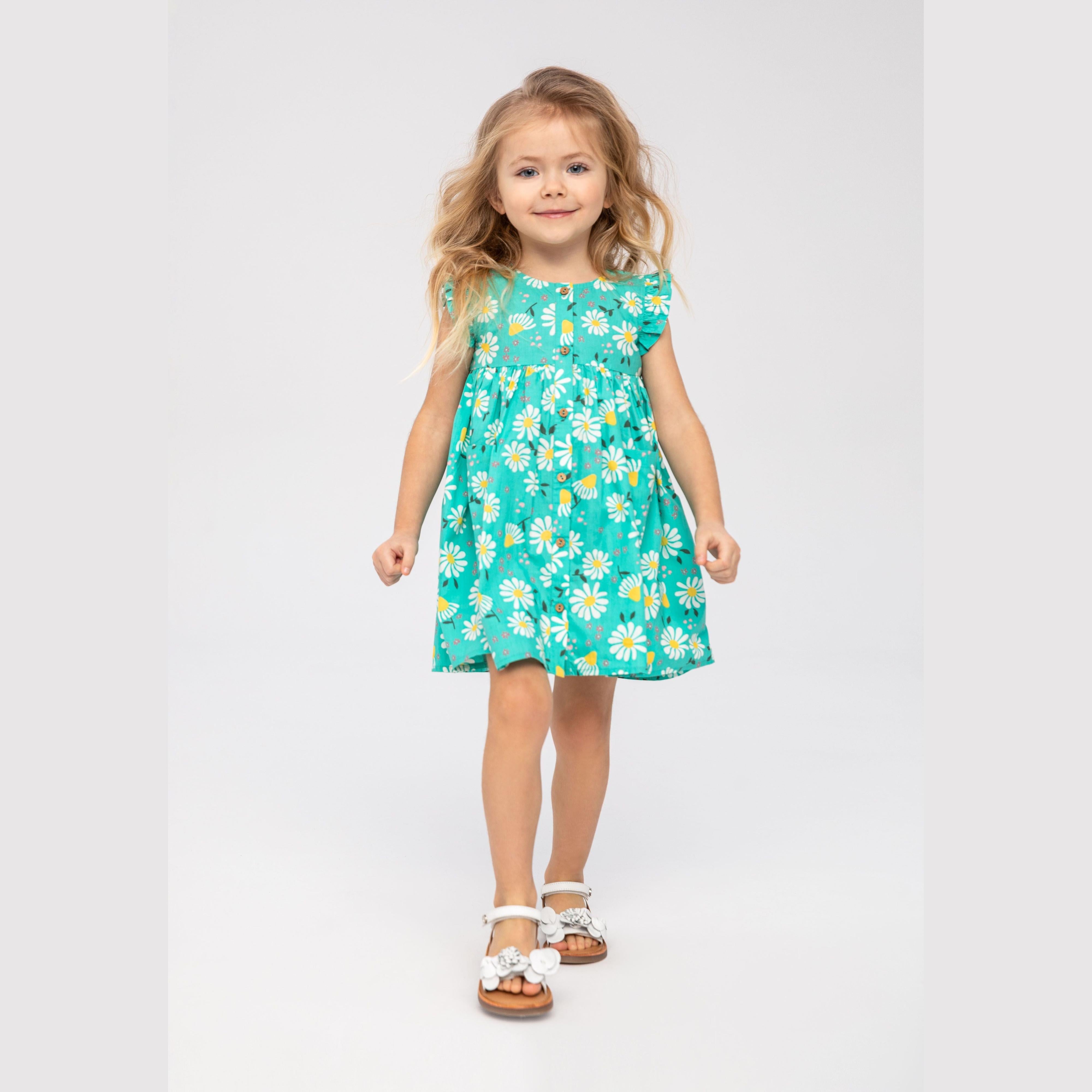 Young blonde girl wearing turquoise dress with white daisy print and frilled sleeves, standing against white background with white sandals