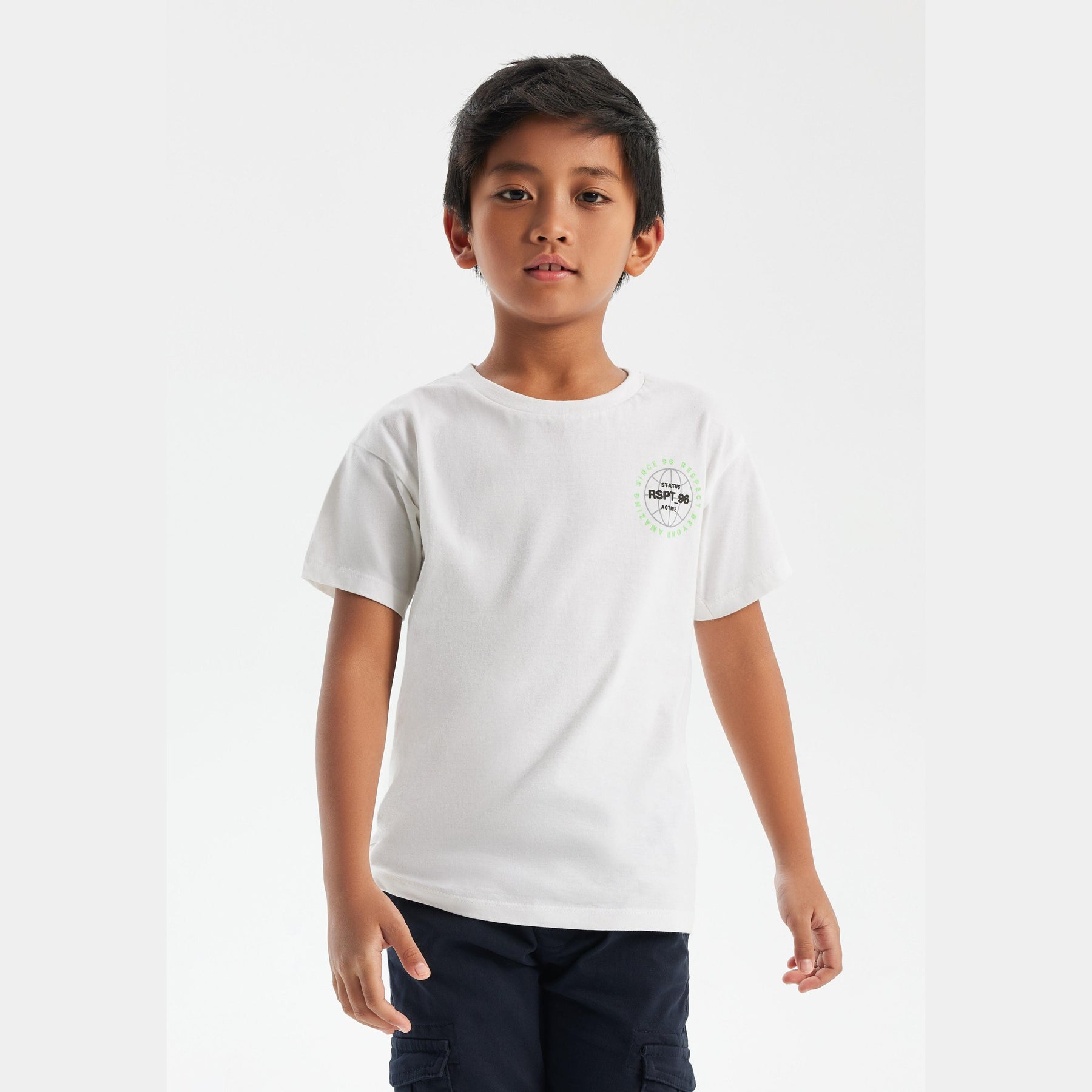 Young boy wearing white t-shirt with small chest logo, paired with navy cargo pants, standing against light grey background