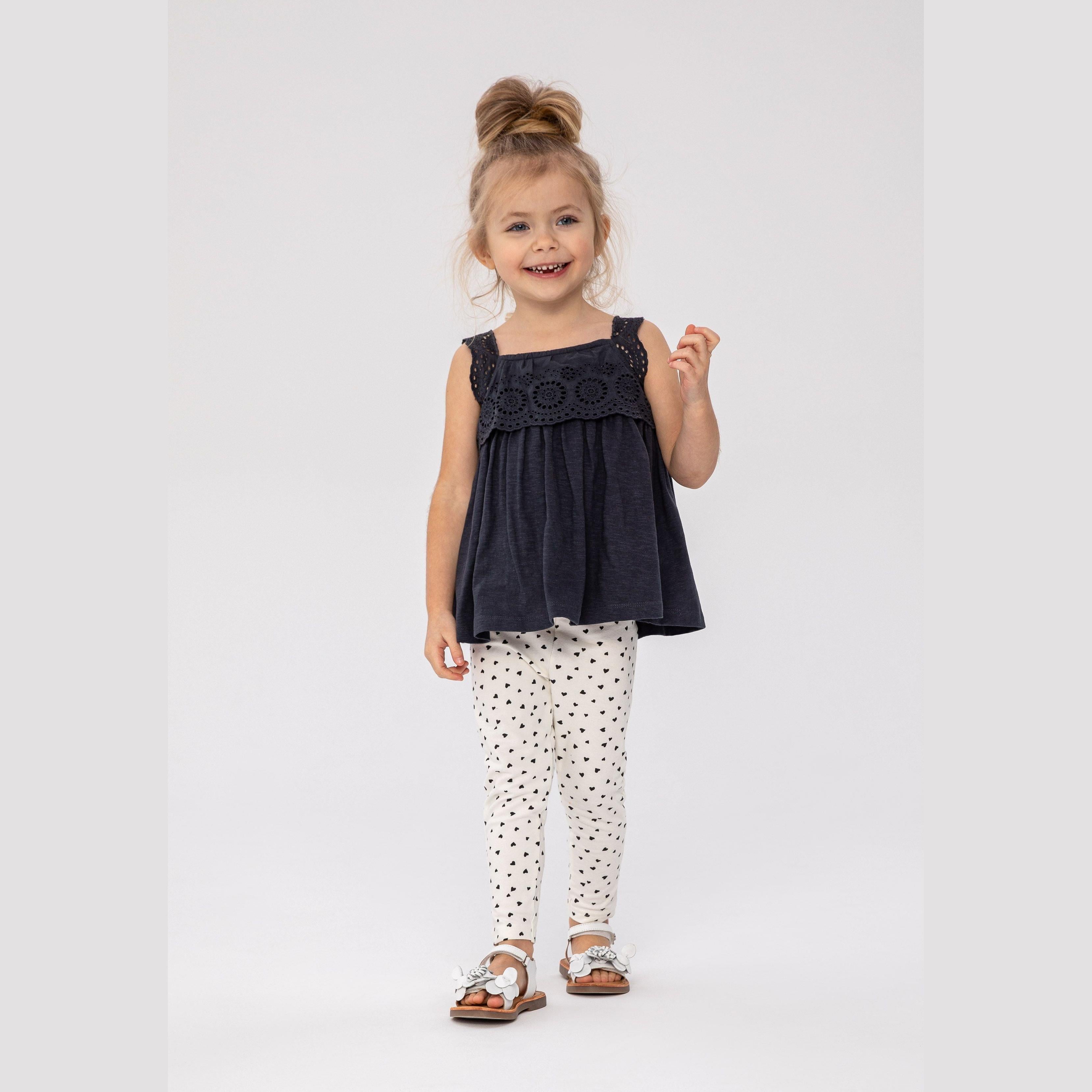 Young girl wearing grey broderie anglaise sleeveless top with ruffle detail and cream heart-print leggings, standing against white background