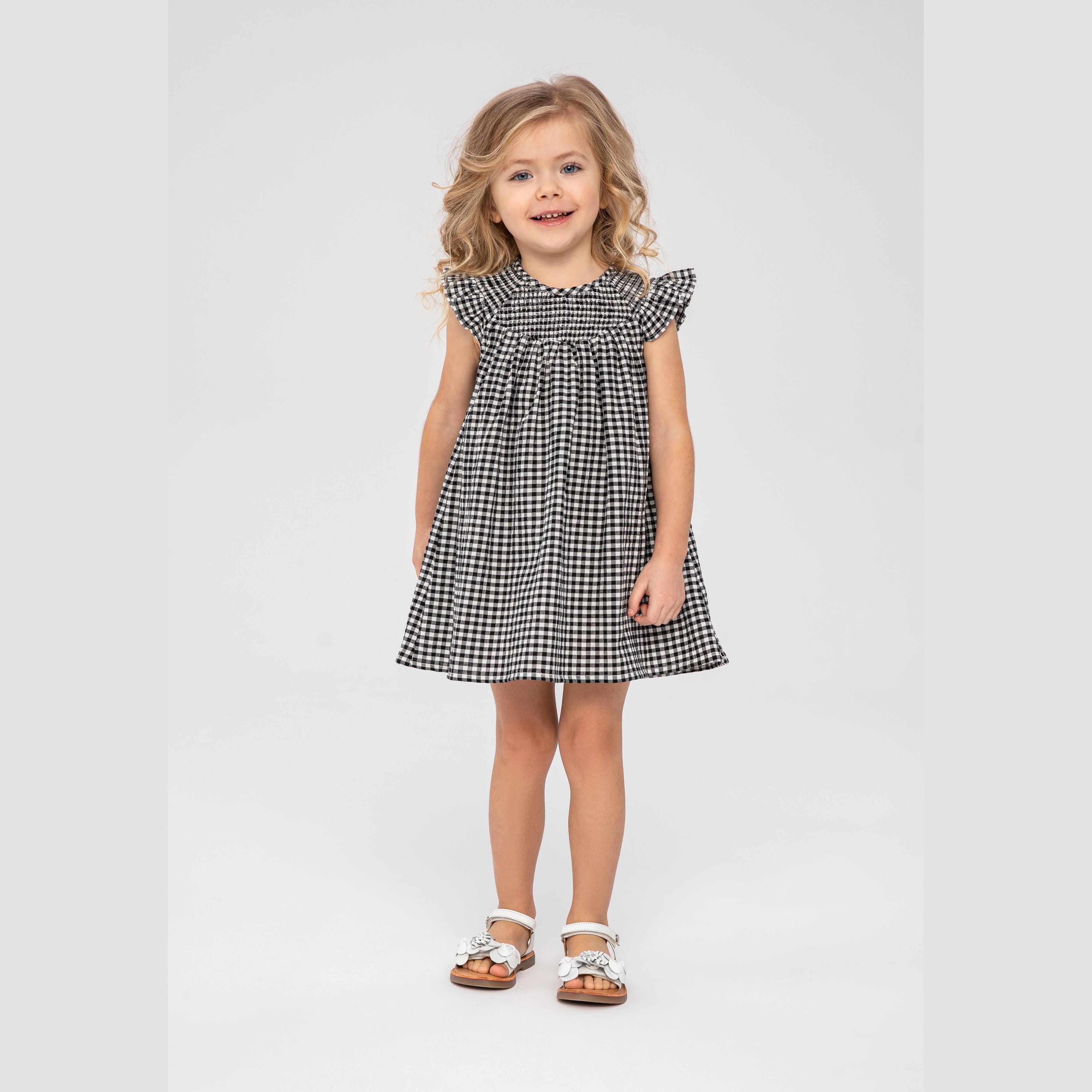 Young girl wearing grey gingham smock dress with frill sleeves, standing against white background, full body view showing dress length and styling