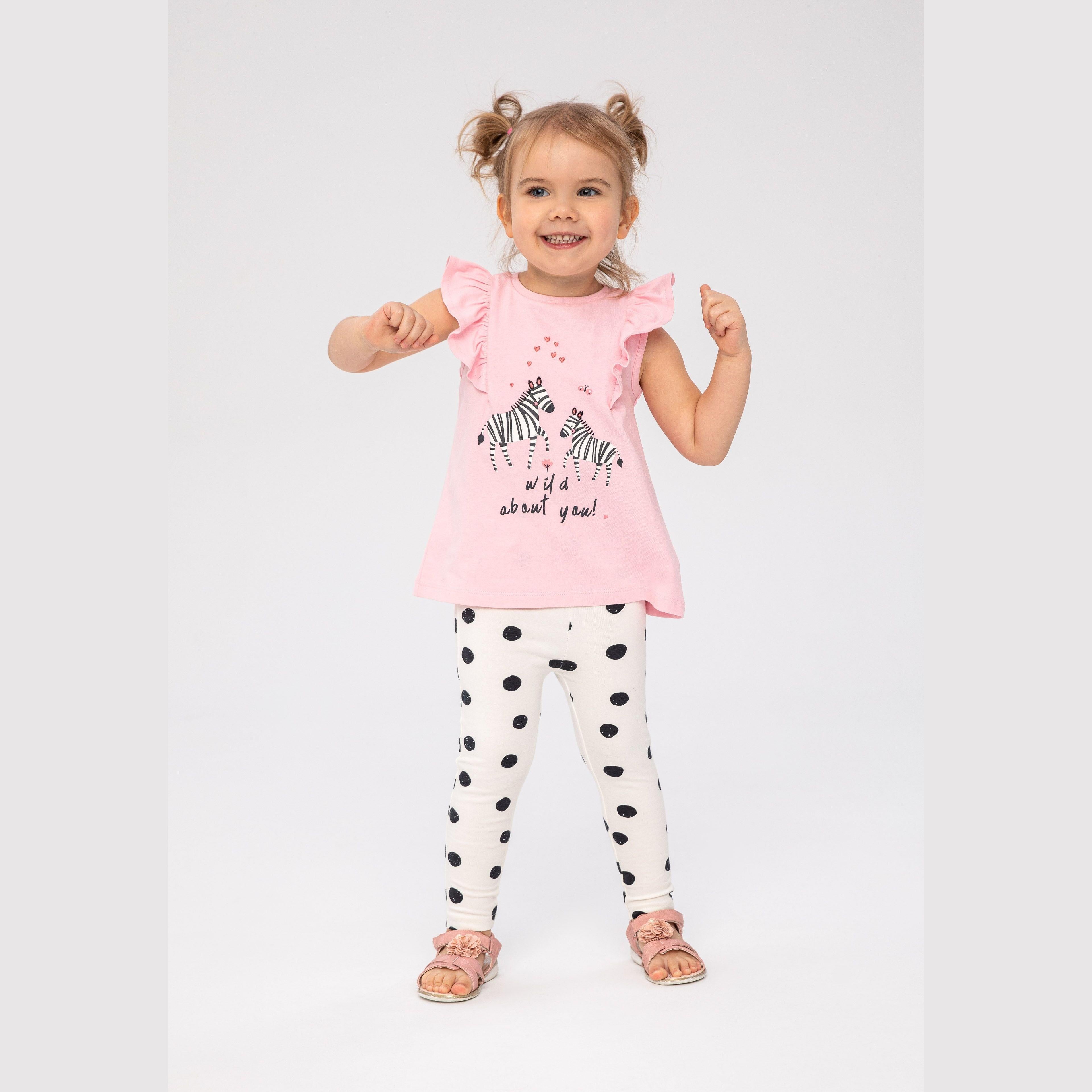 Young girl wearing pink zebra print flutter sleeve top with white polka dot leggings, standing against white studio background