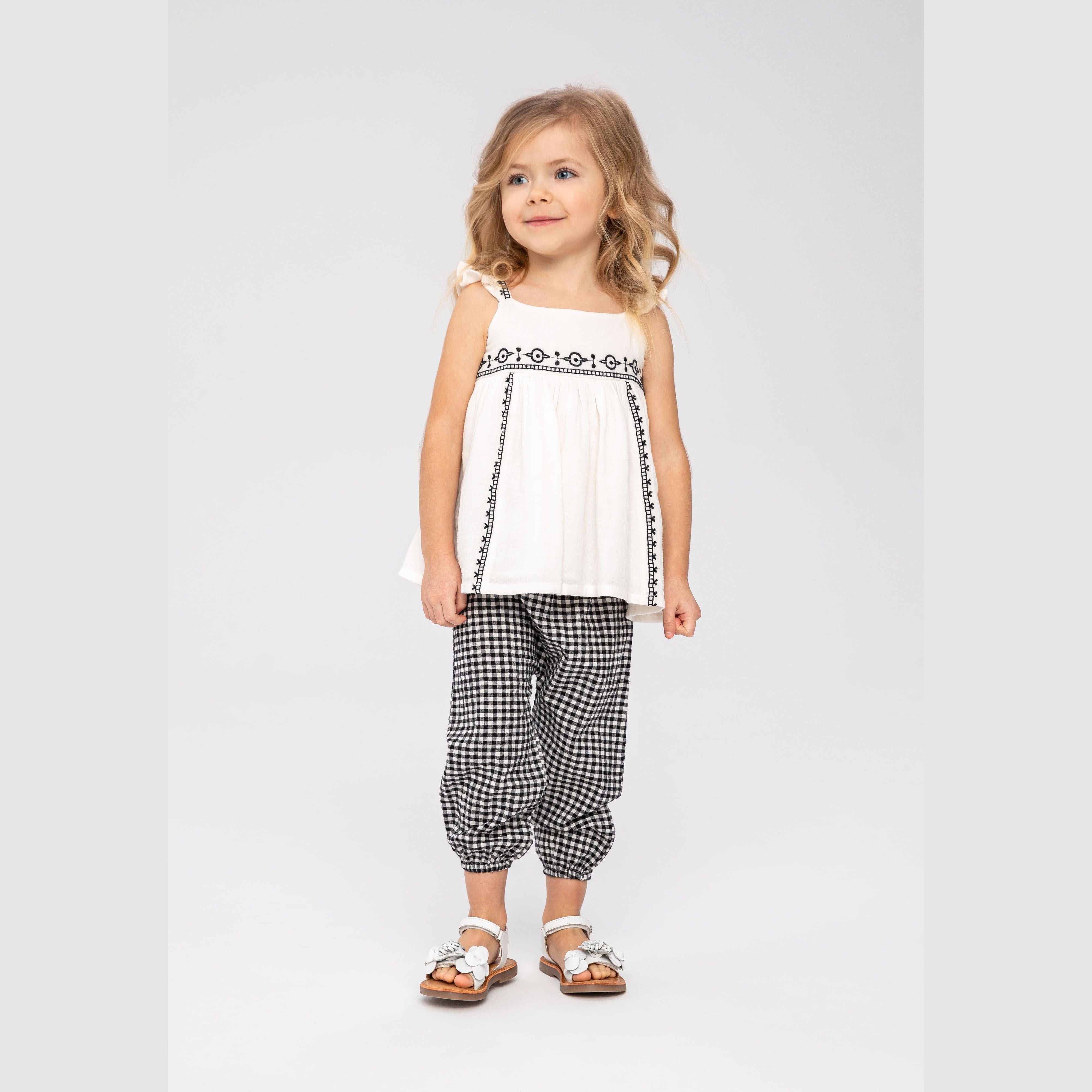 Young girl wearing cream embroidered sleeveless top with frill straps and black gingham harem trousers, standing against light grey background