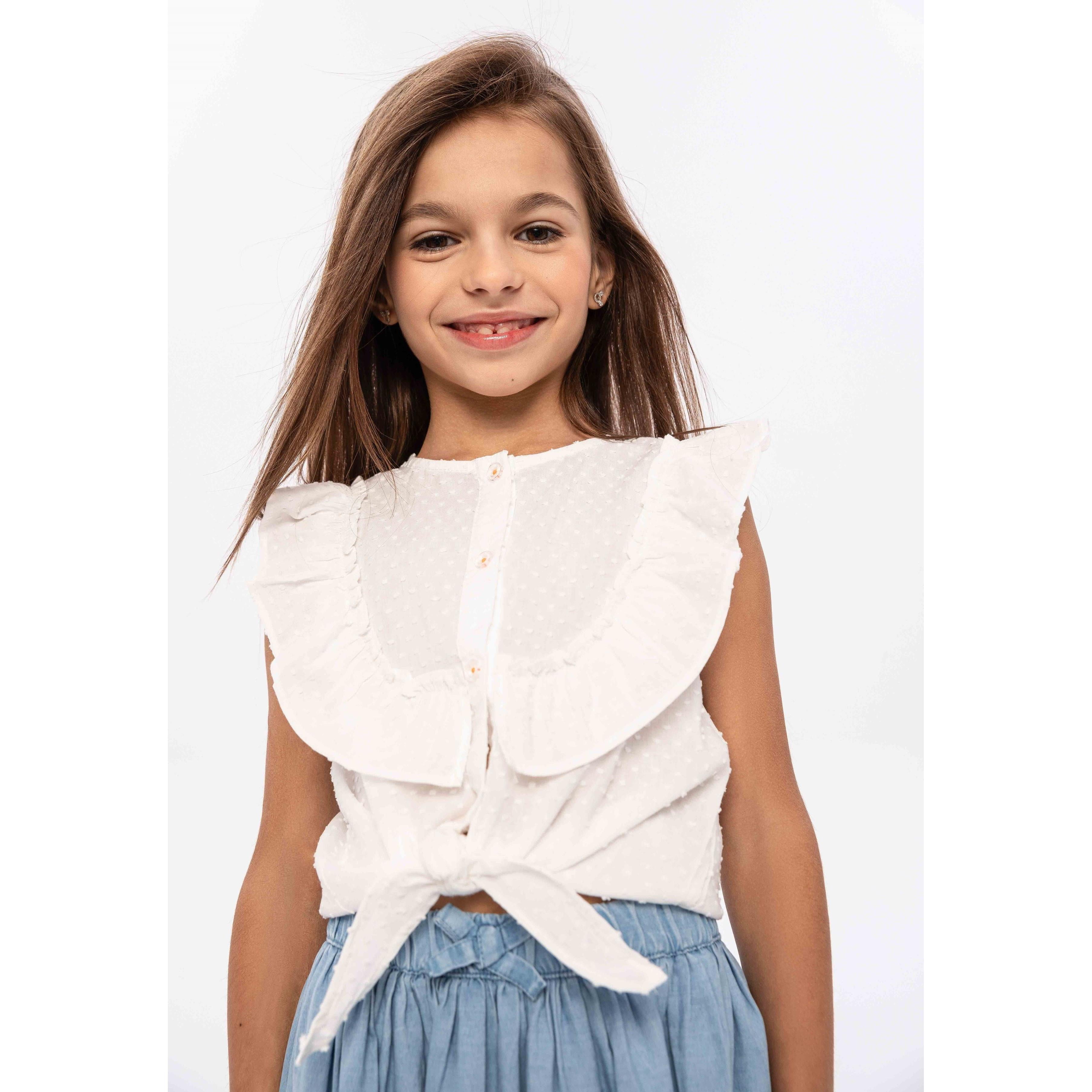Young girl wearing white broderie anglais sleeveless top with ruffle collar and tie front, paired with denim skirt, smiling against white background