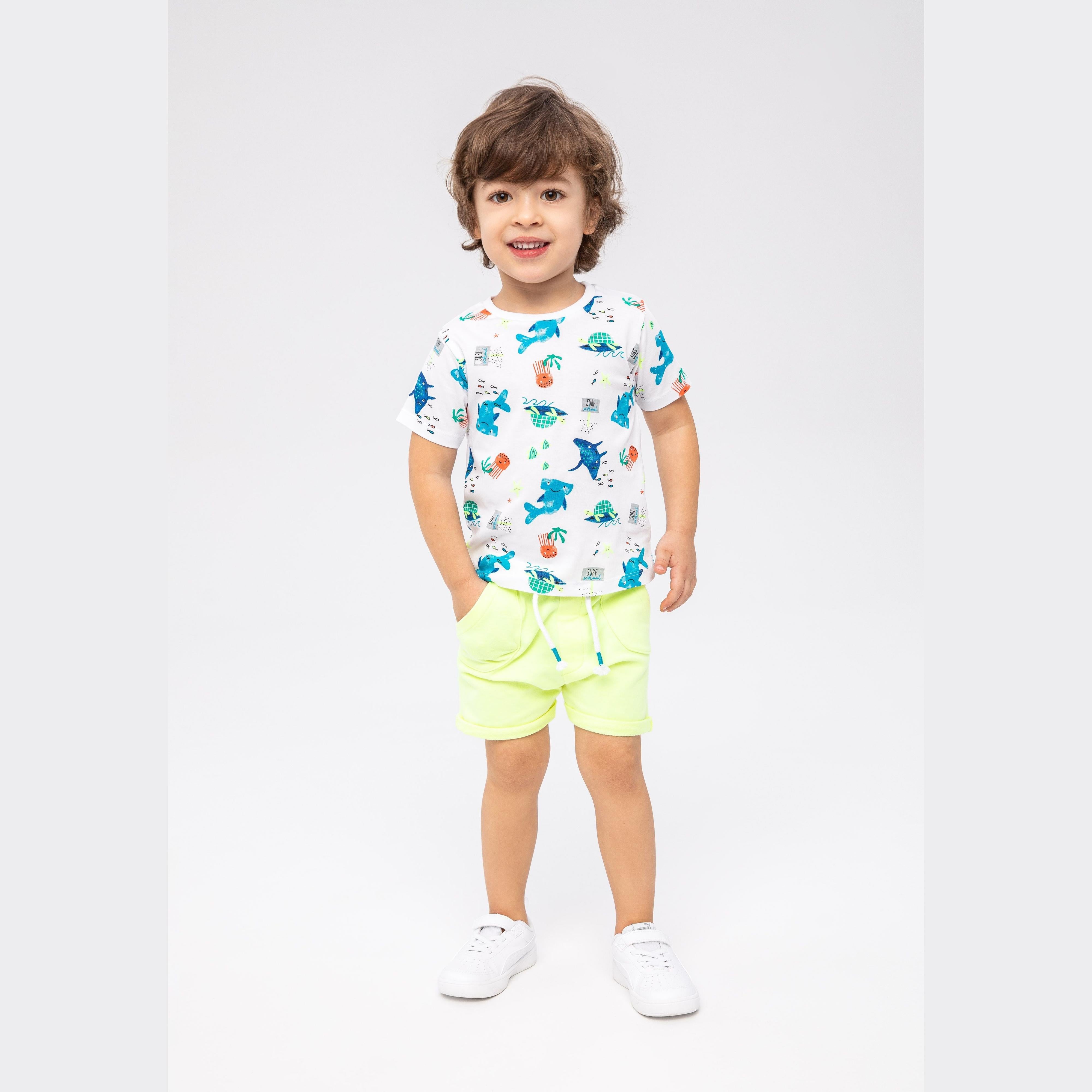 Young boy wearing white shark print t-shirt and lime green shorts, smiling at camera against light grey background, full body shot
