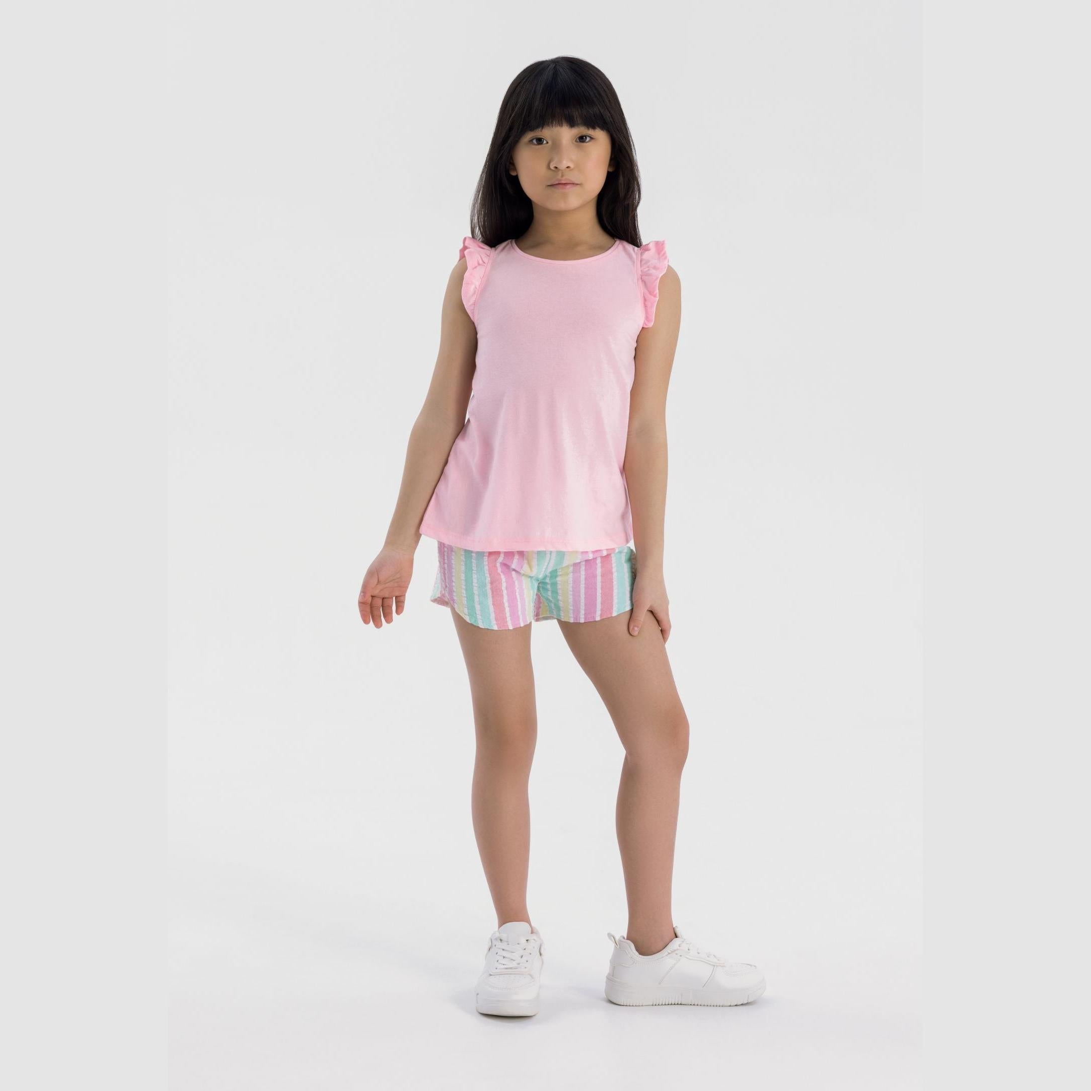 Young girl wearing pink sleeveless top with ruffled shoulders and multicoloured striped shorts, standing against white background
