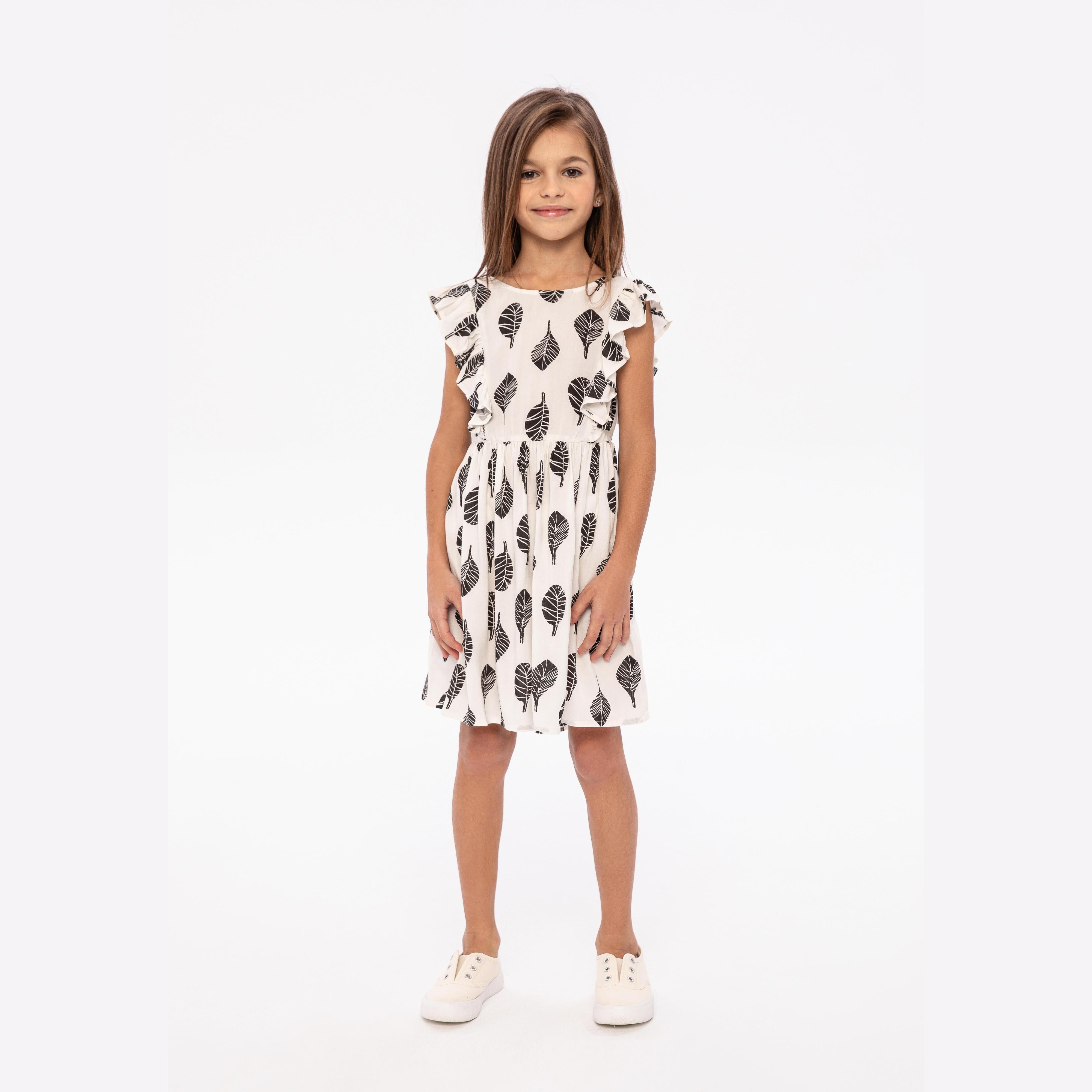 Young girl wearing white summer dress with black leaf print and frill sleeves, standing against white background