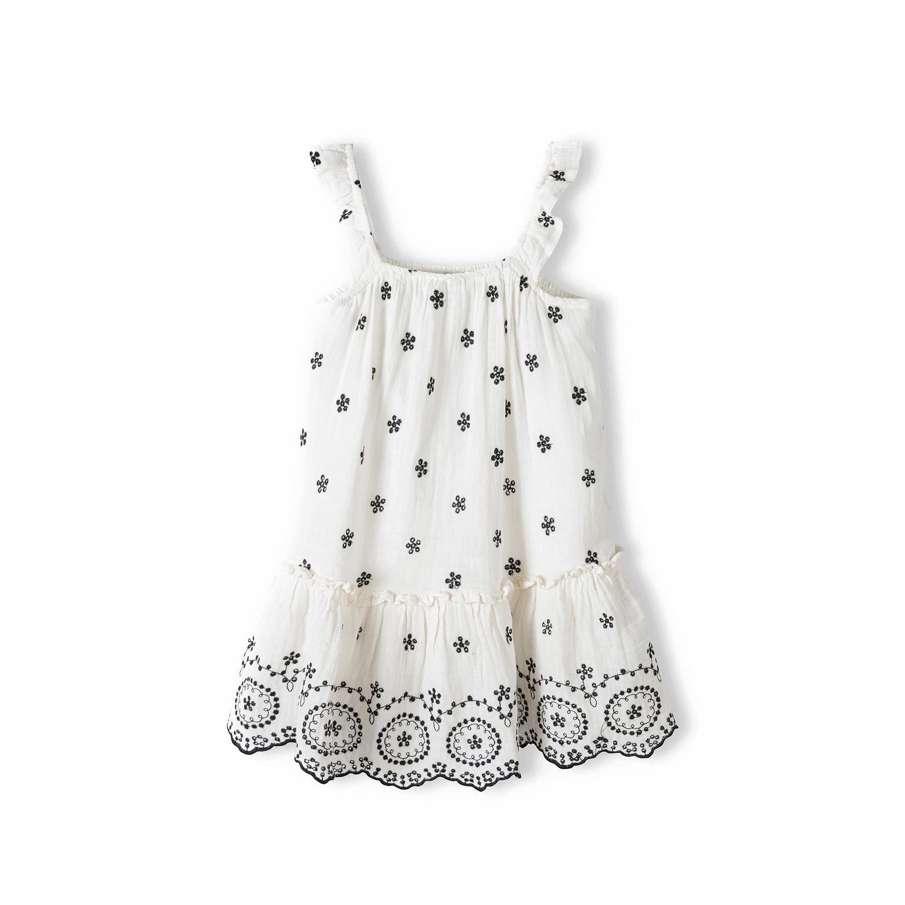 White embroidered dress laid flat showing black floral motifs throughout fabric, frilled shoulder straps and scalloped embroidered hem detail
