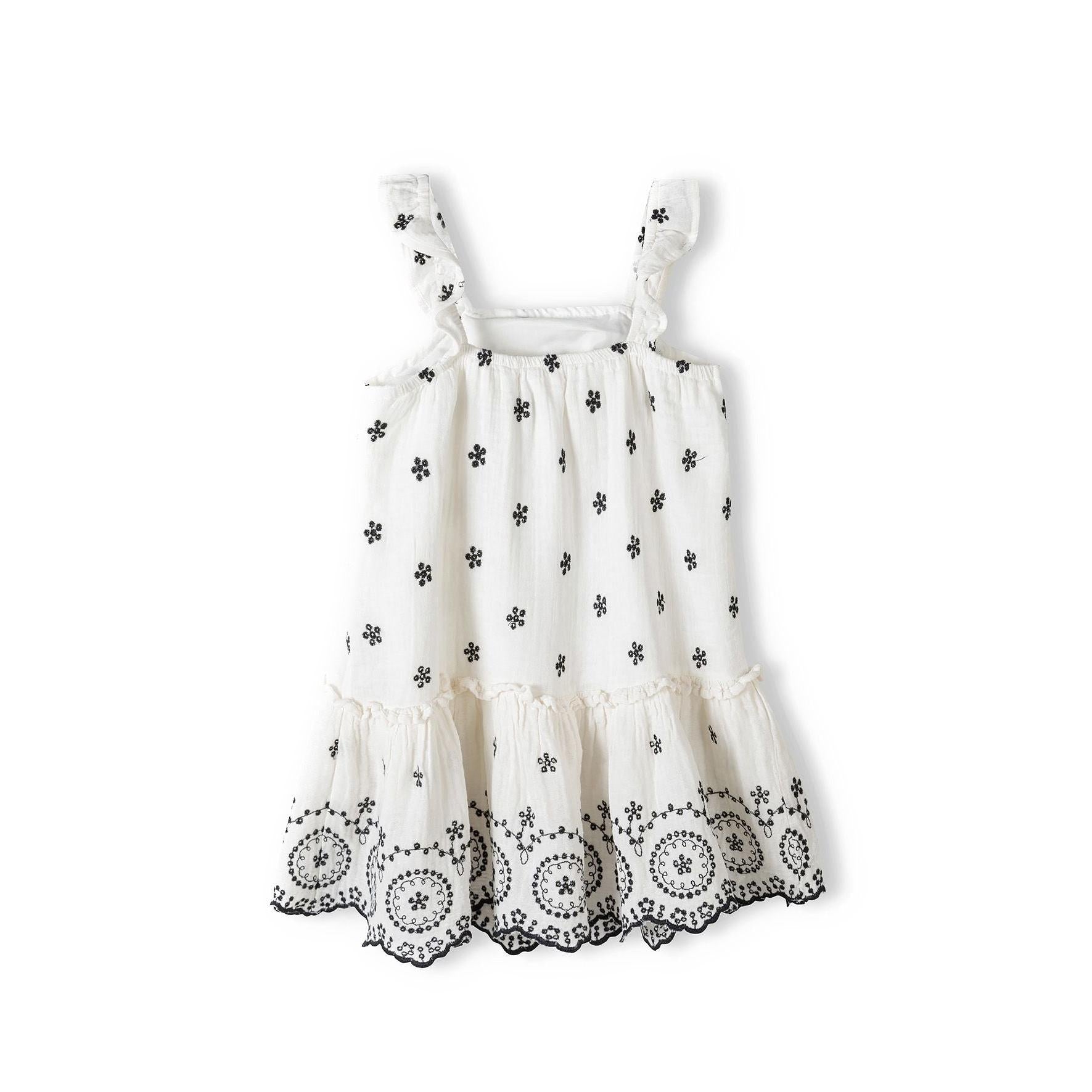 White embroidered dress laid flat showing black floral motifs throughout fabric, frilled shoulder straps and scalloped embroidered hem detail