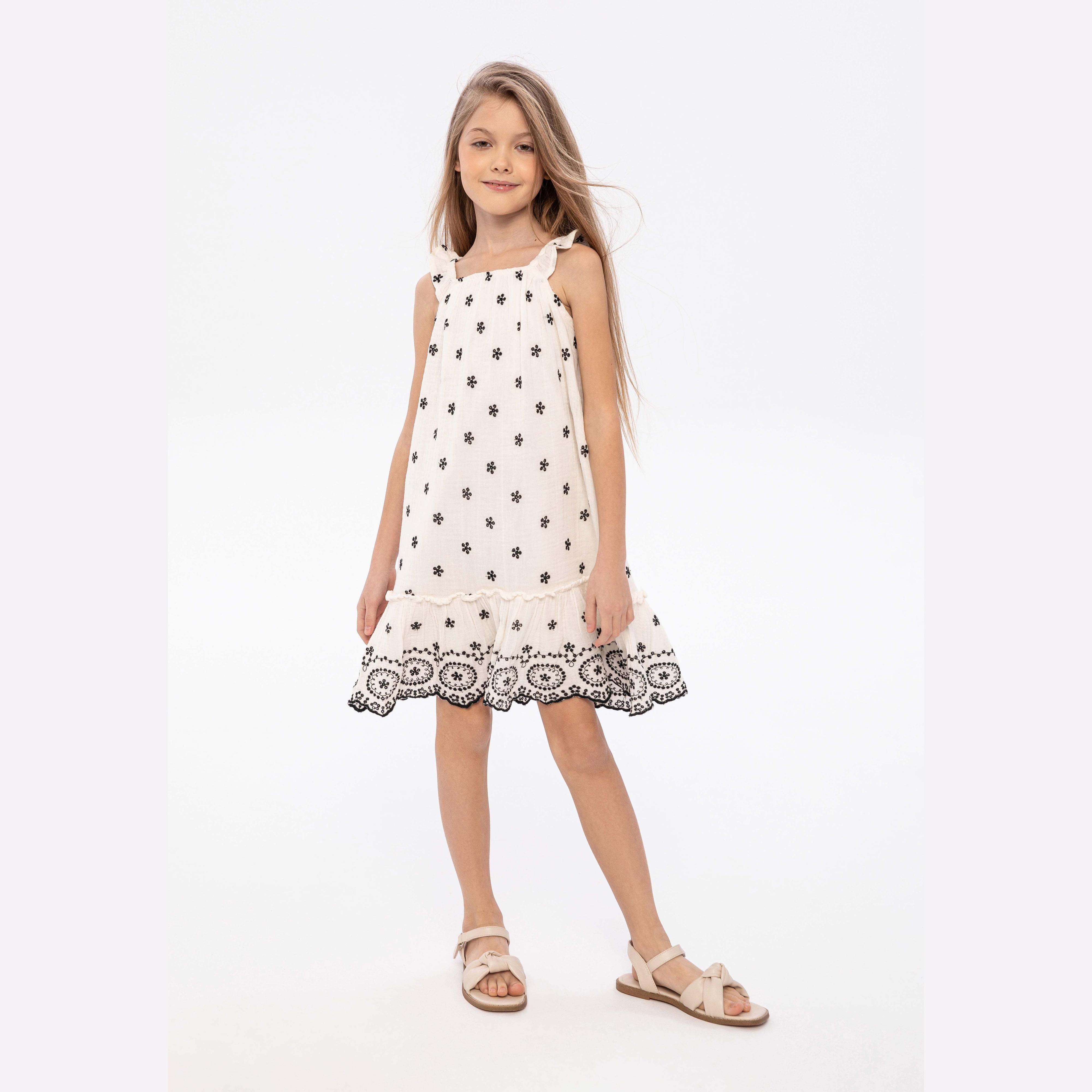 Young girl wearing white embroidered dress with black floral motifs and frilled straps, standing against light grey background wearing beige sandals