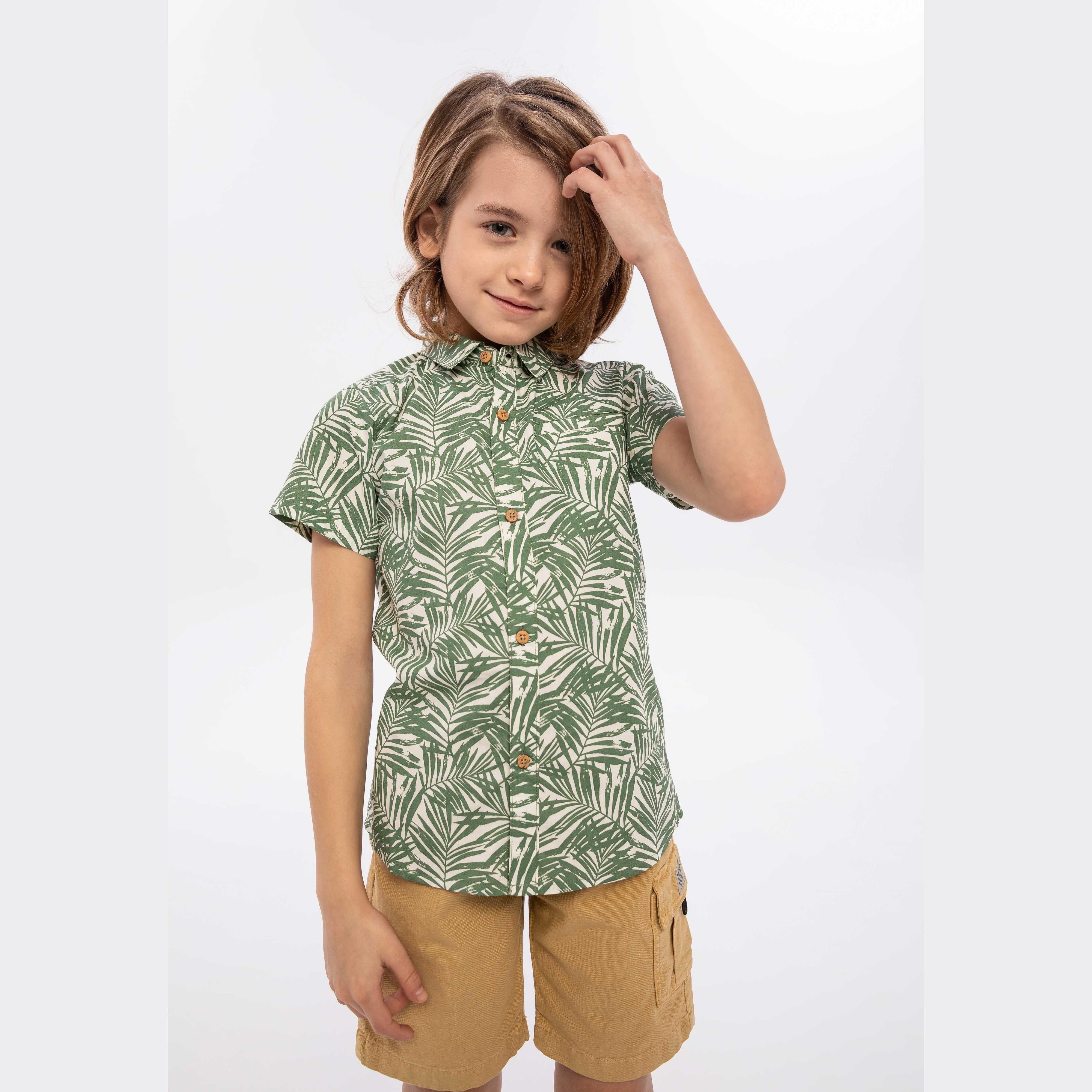 Young boy wearing olive green palm print short sleeve shirt with natural wooden buttons, paired with tan corduroy shorts, against white studio background