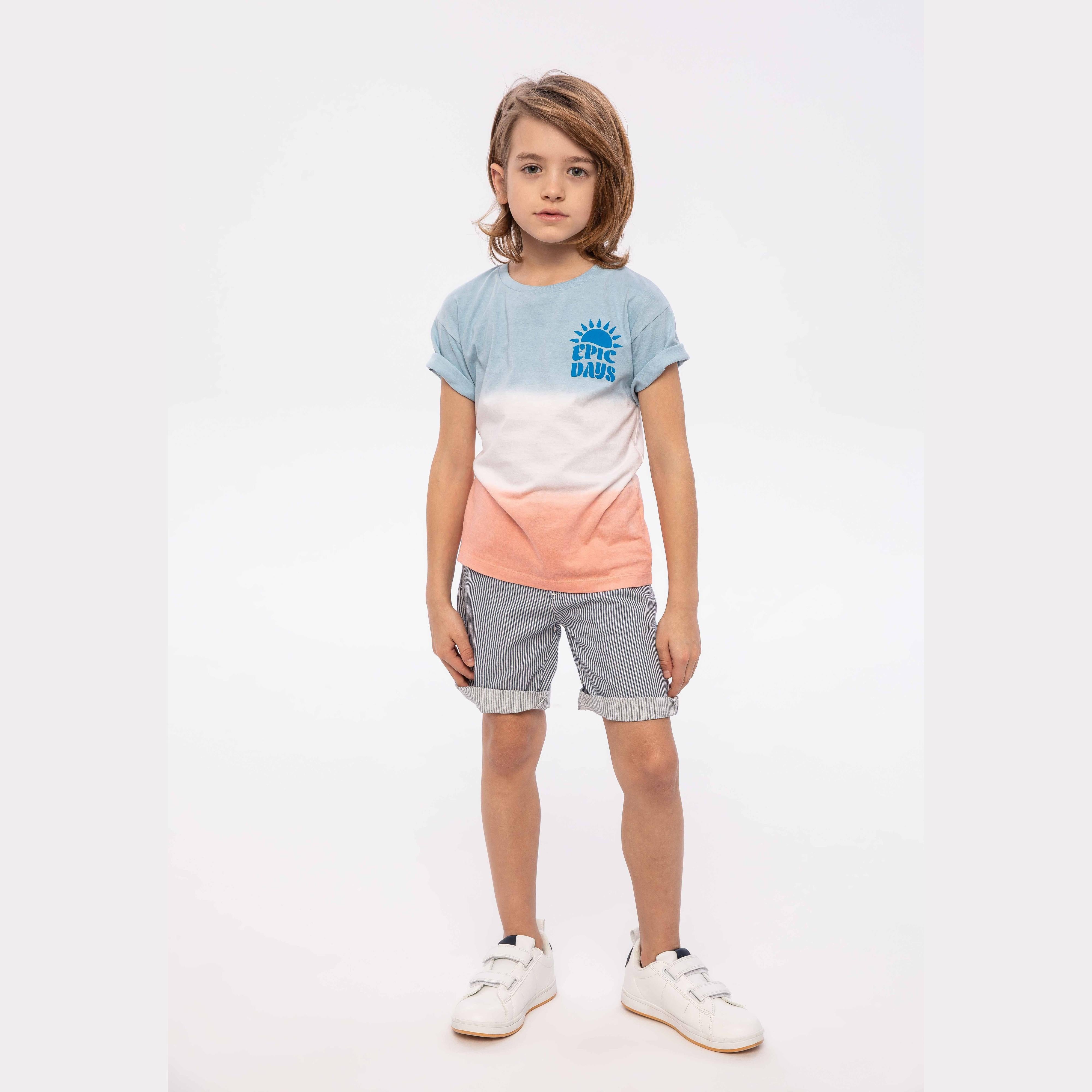 Young child wearing light blue to coral dip dye t-shirt with Epic Days sun print and striped shorts, standing against white studio background