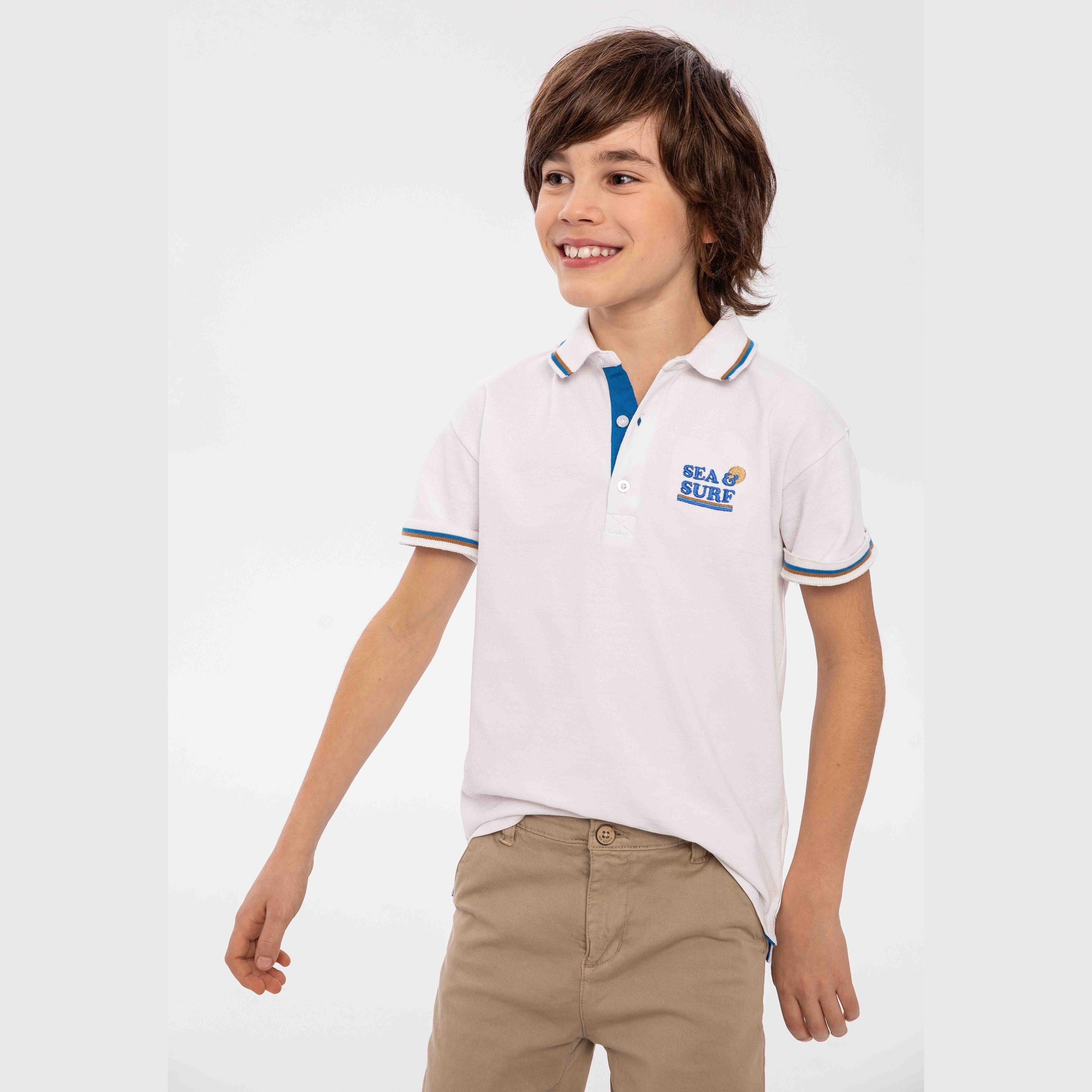 Boy wearing white polo shirt with contrast tipping and beige chinos, front view on light grey background