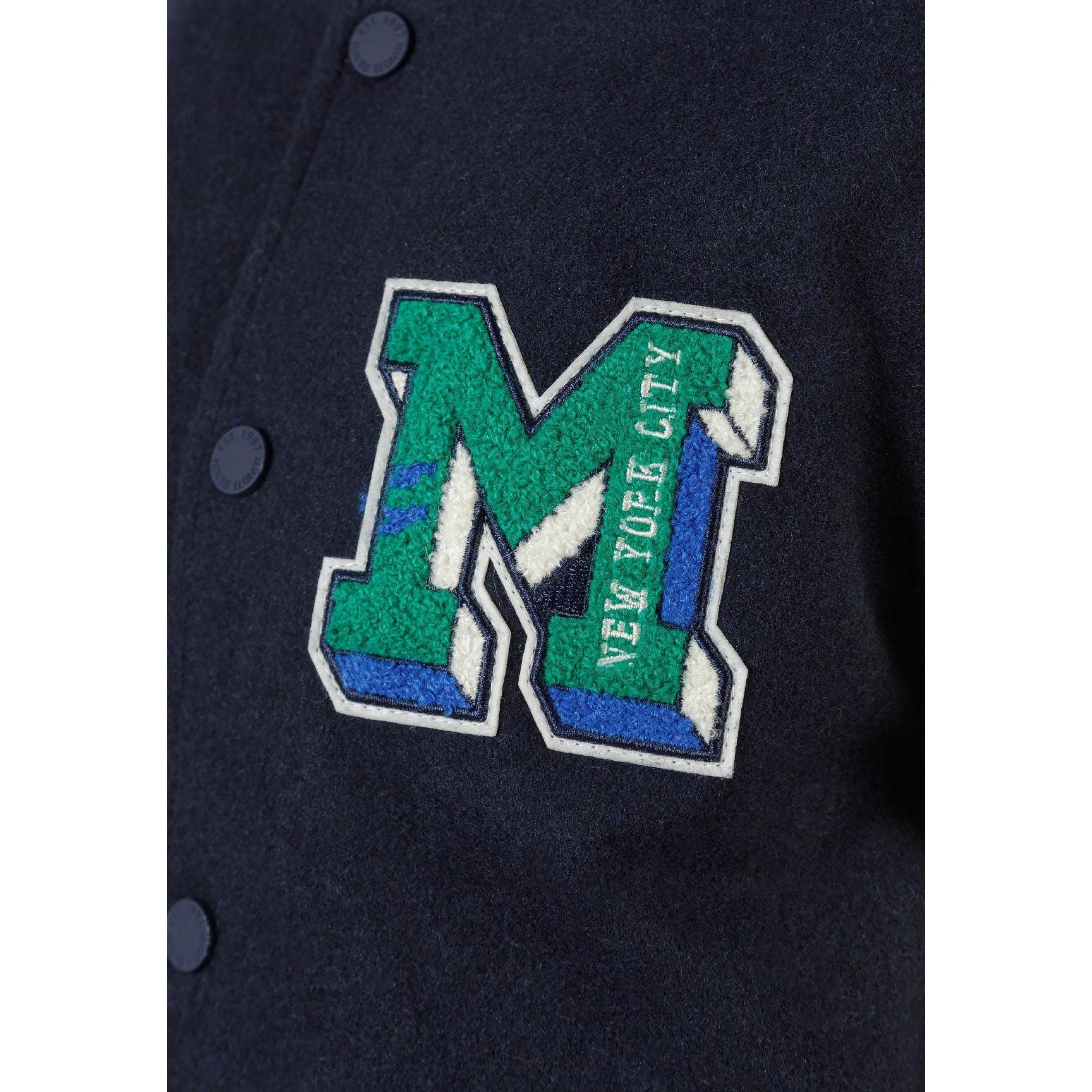 Close-up detail of green chenille M appliqué patch with NEW YORK CITY text on navy fabric background with press studs visible