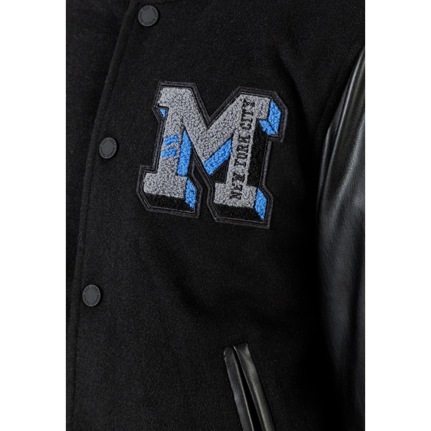 Close-up detail of appliqué M logo in grey and blue with text, showing press stud fastening and black felt fabric texture