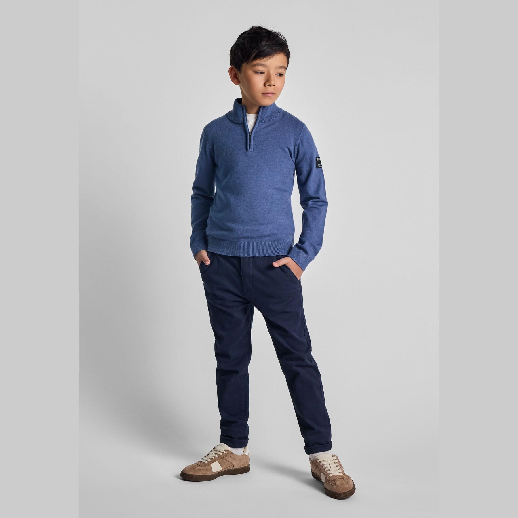 Boy wearing navy chino trousers with blue quarter-zip sweater standing against white background