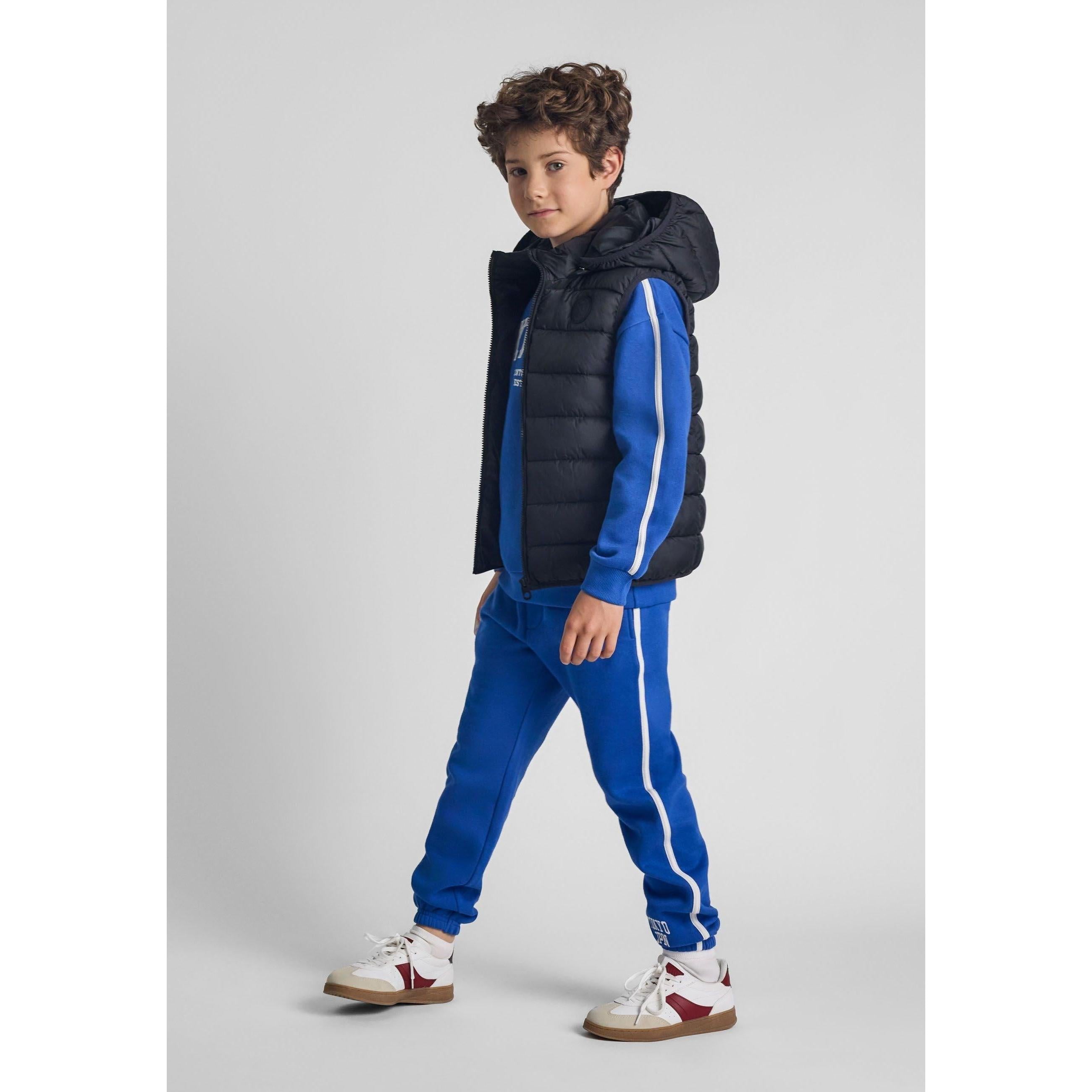 Young boy wearing black padded gilet over blue tracksuit, standing against grey background, showing full outfit styling
