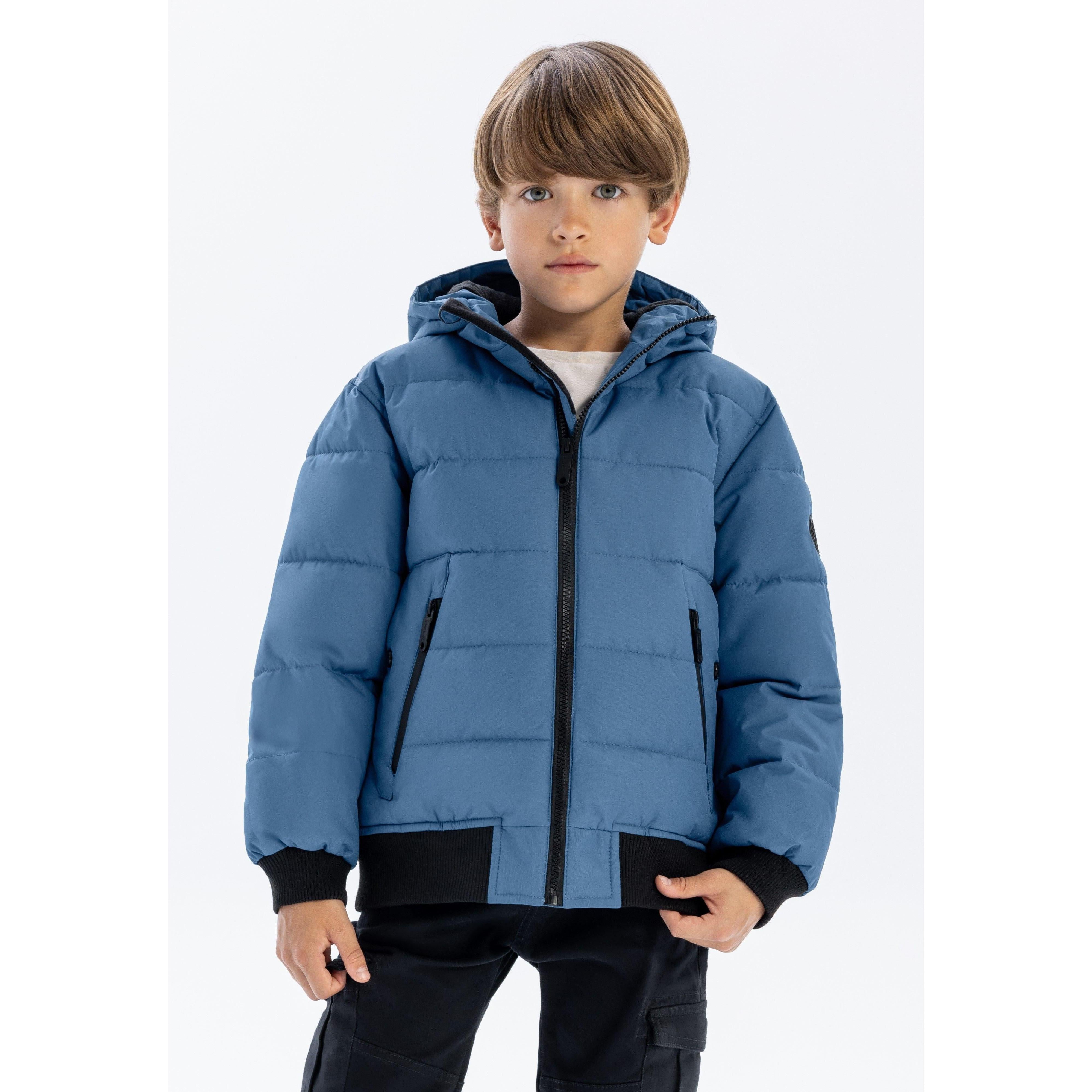 Young boy wearing blue padded jacket with hood and zip pockets, standing against white background