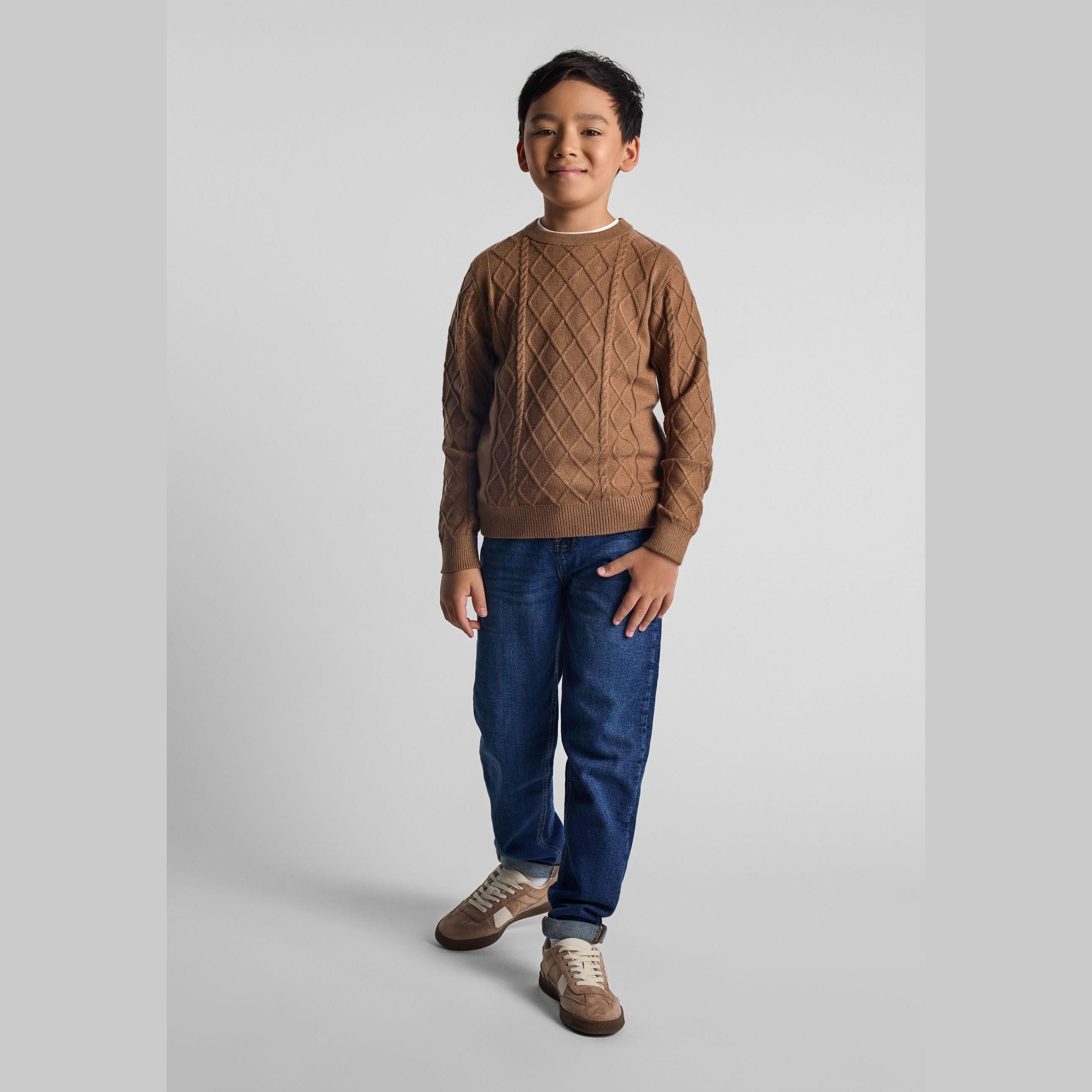 Young boy wearing brown cable knit jumper with diamond pattern and blue jeans, standing against white studio background