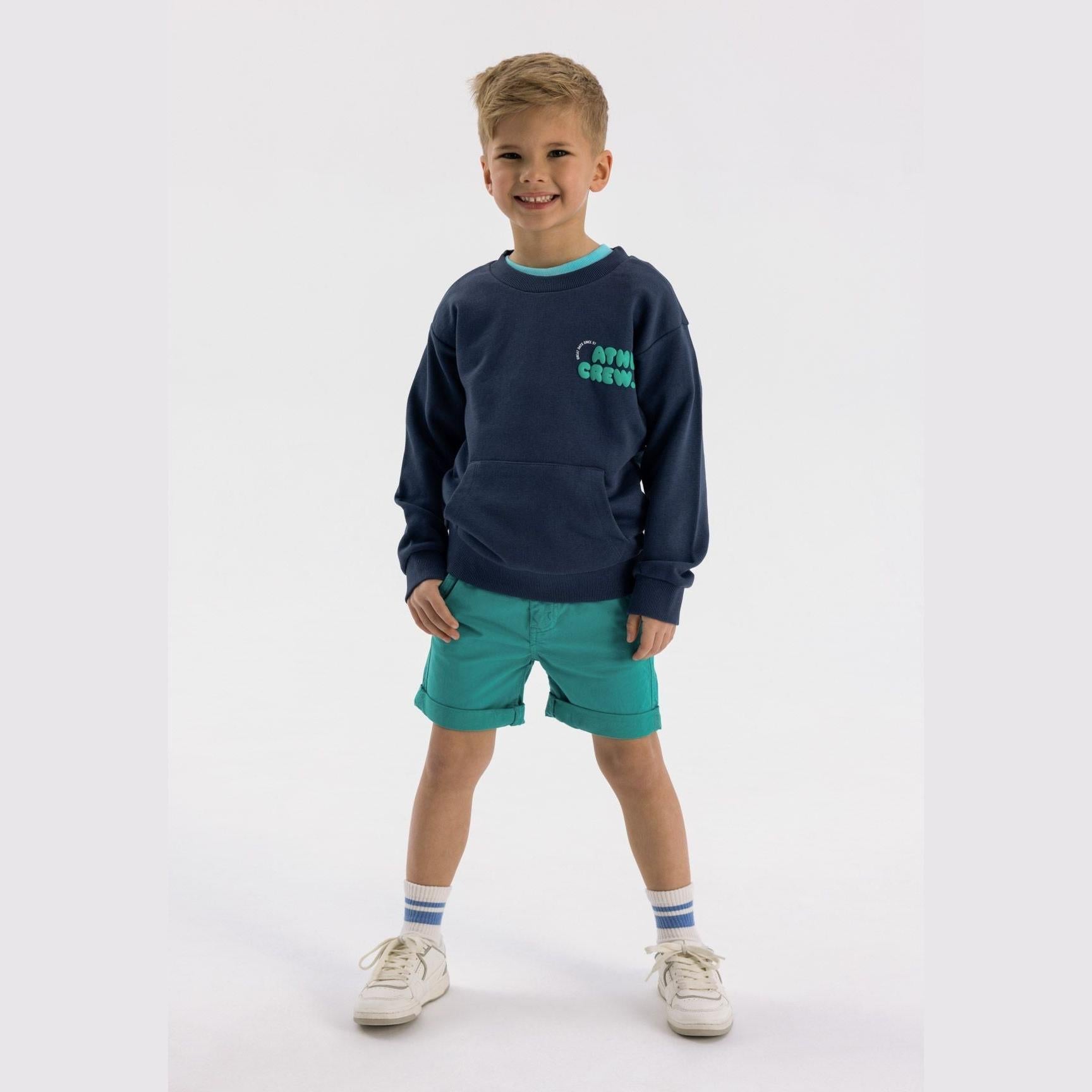 Young blonde boy wearing navy crew neck sweatshirt with teal shorts, white striped socks and cream trainers against white background
