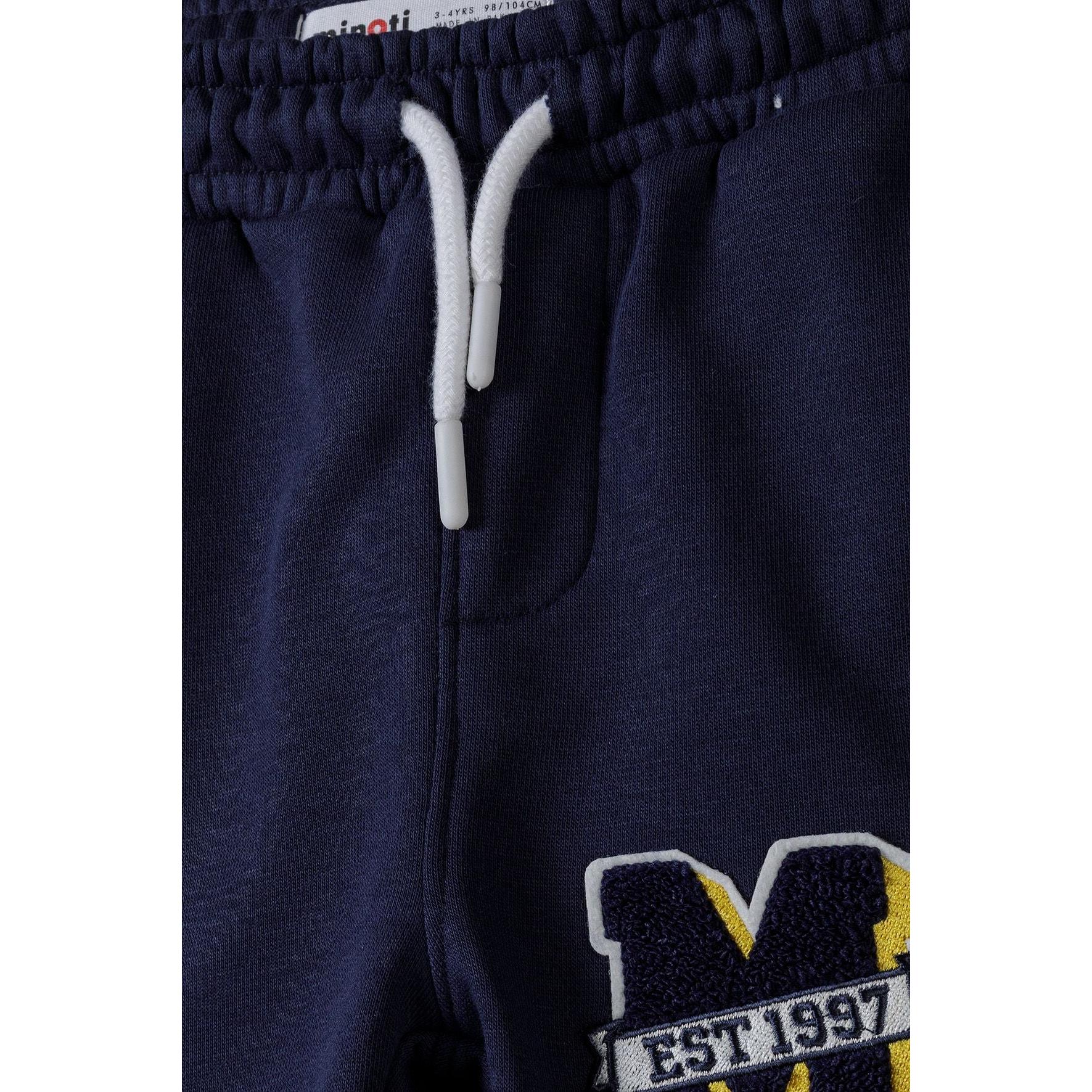 Close-up detail of navy sweatpants waistband showing white drawstring ties and embroidered M EST 1997 logo patch with yellow and white stitching