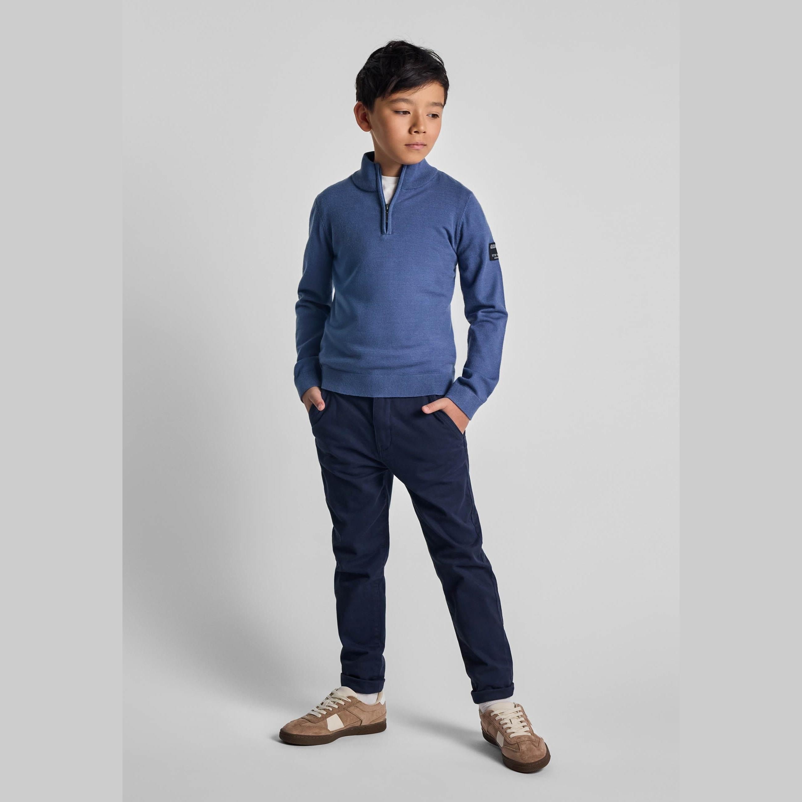 Boy wearing blue half zip jumper with stand collar and dark navy trousers, standing against light grey background