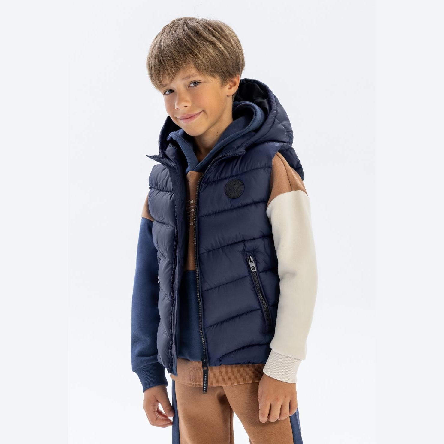 Boy wearing navy padded gilet with detachable hood over cream sweater and brown trousers on white background