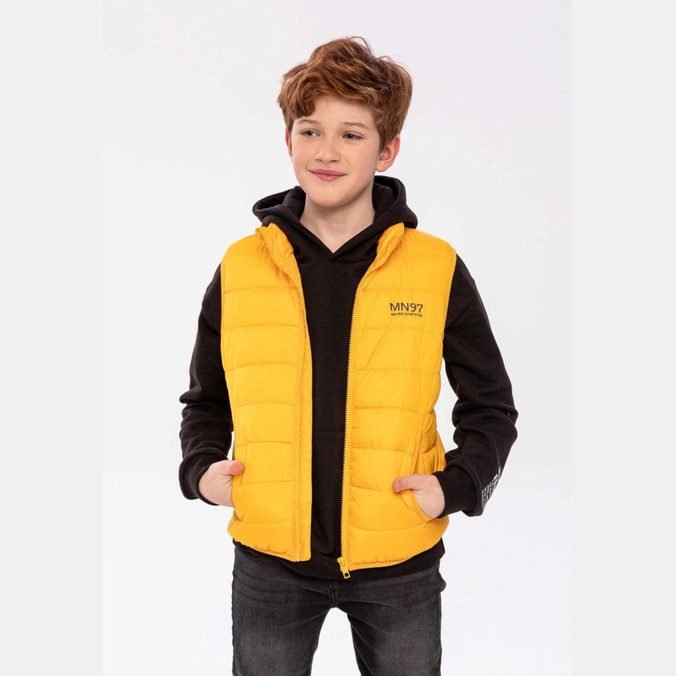 Boy wearing bright yellow padded gilet over black hoodie and dark jeans, standing against white studio background