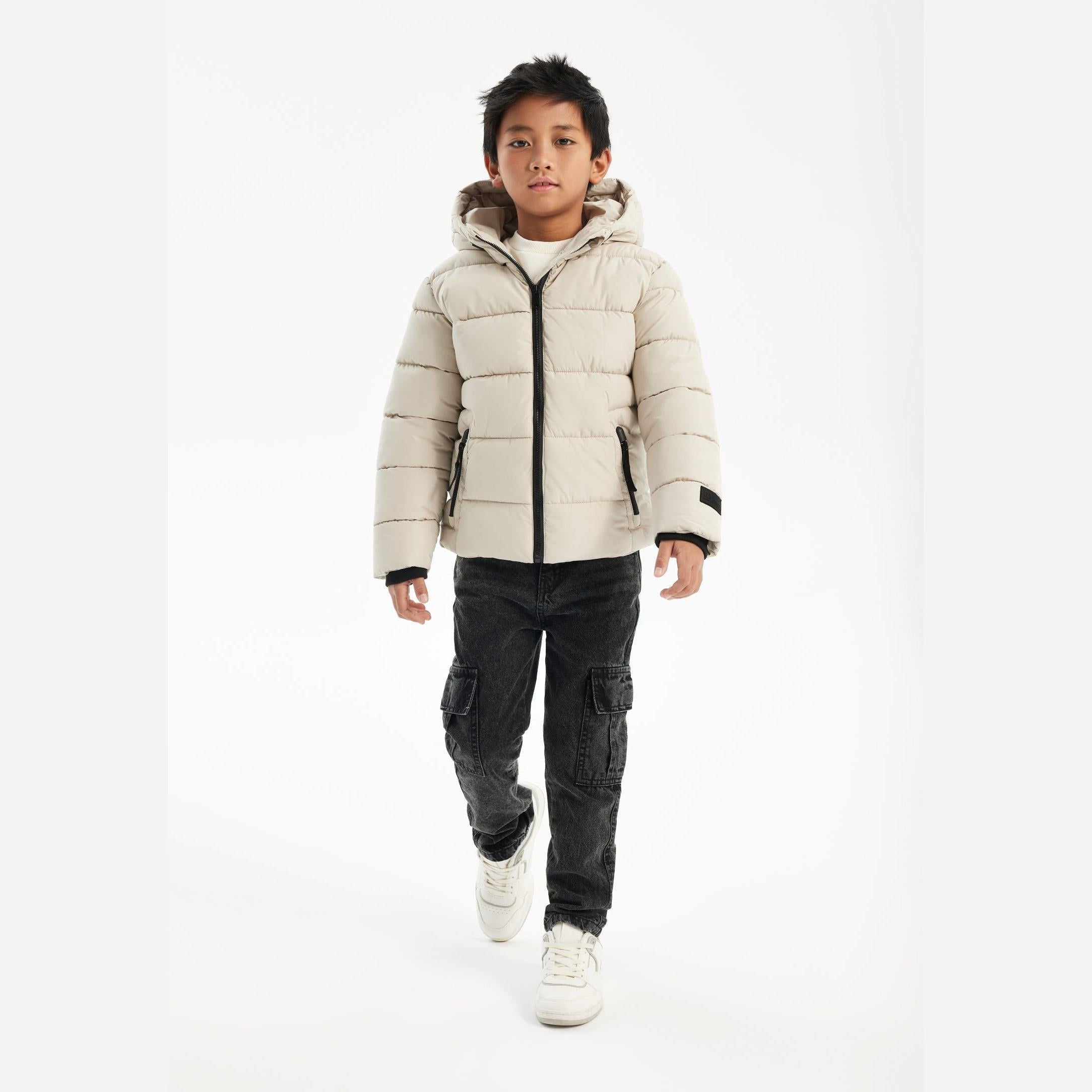 Young boy wearing cream padded jacket with hood and dark cargo pants, standing against white background with hands at sides