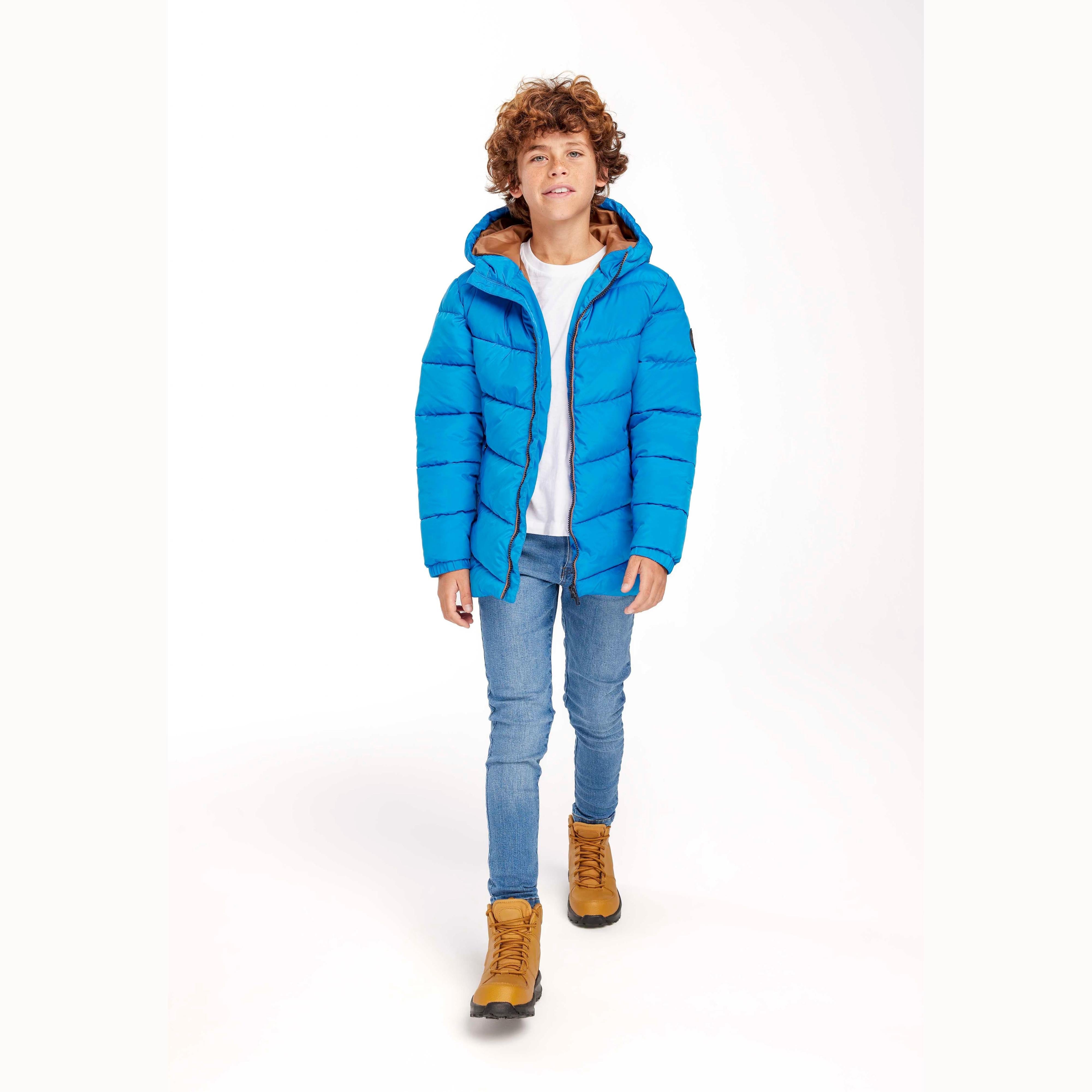 Young boy with curly hair wearing cobalt blue hooded padded jacket over white t-shirt and jeans, standing against white background