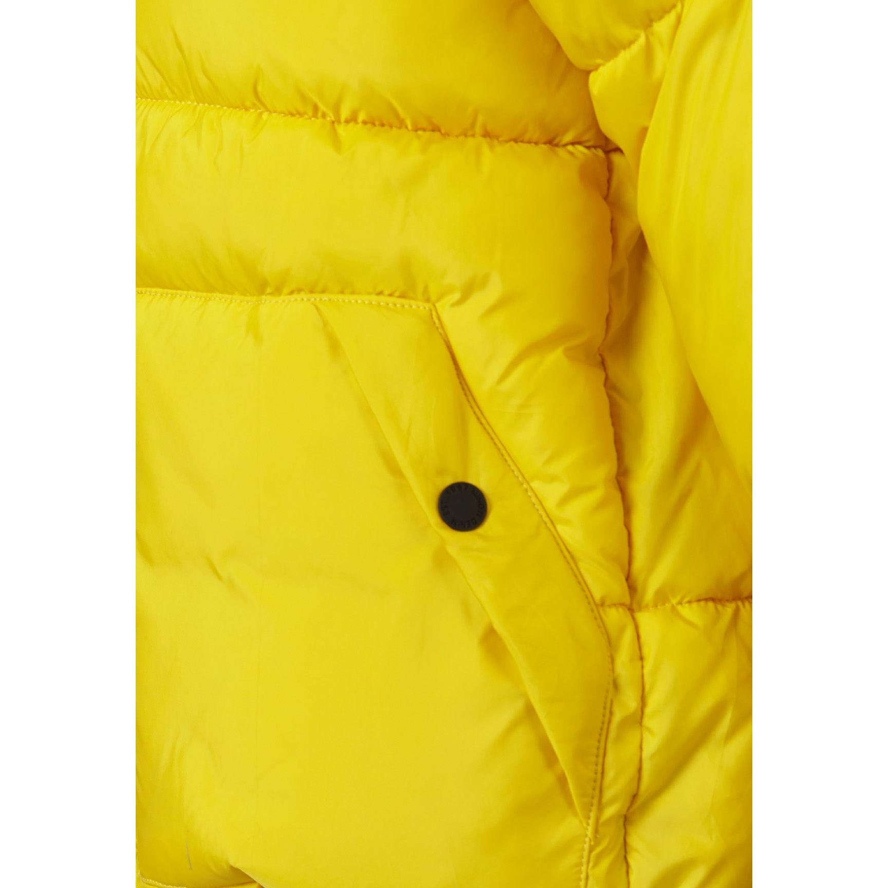 Close-up detail of black circular Minoti logo patch with crossed design on bright yellow padded jacket fabric showing quilted texture