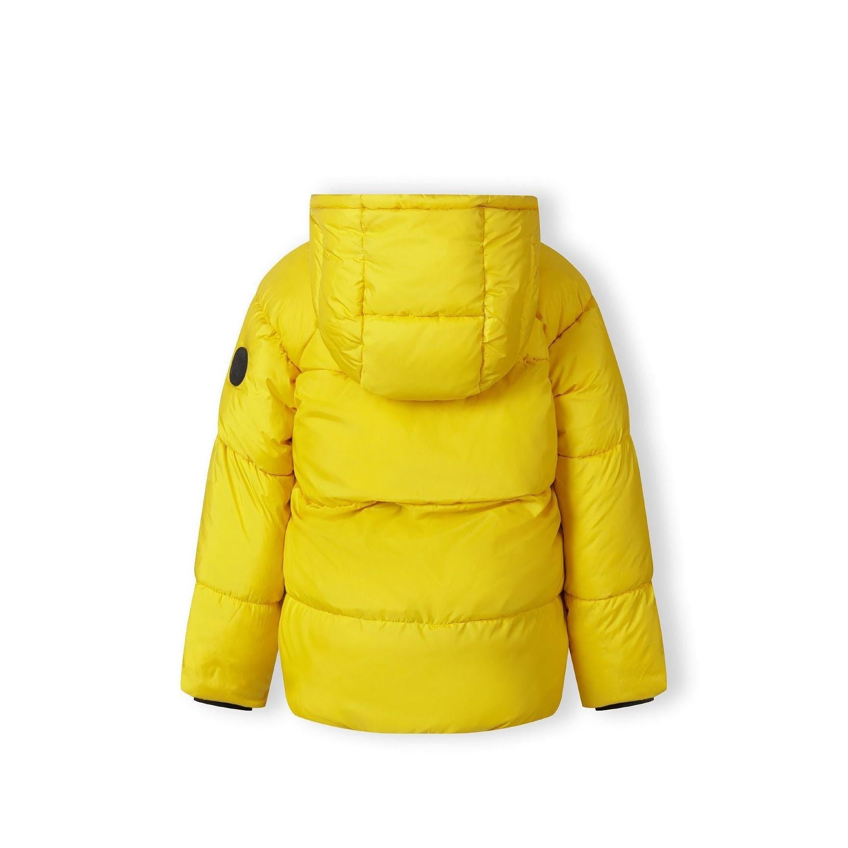 Close-up detail of black circular Minoti logo patch with crossed design on bright yellow padded jacket fabric showing quilted texture